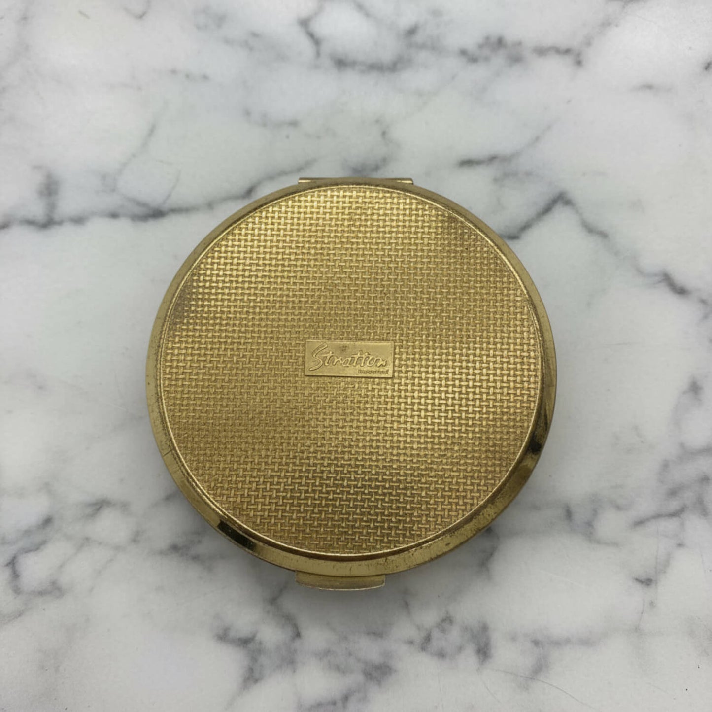 A round gold compact case with a textured grid pattern and a small embossed Sgratton logo in the center, resting on a white and gray marble surface.