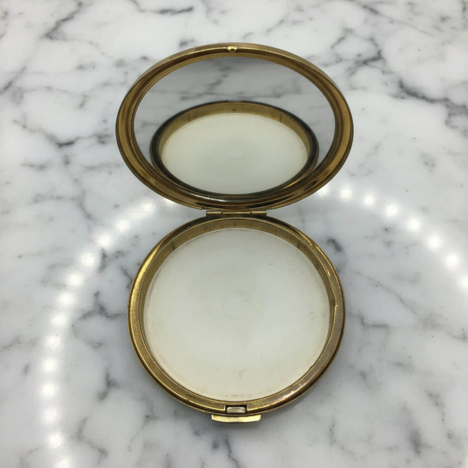 An open round compact mirror with a gold-toned metal frame and a white pressed powder pan, resting on a white and grey marble surface.