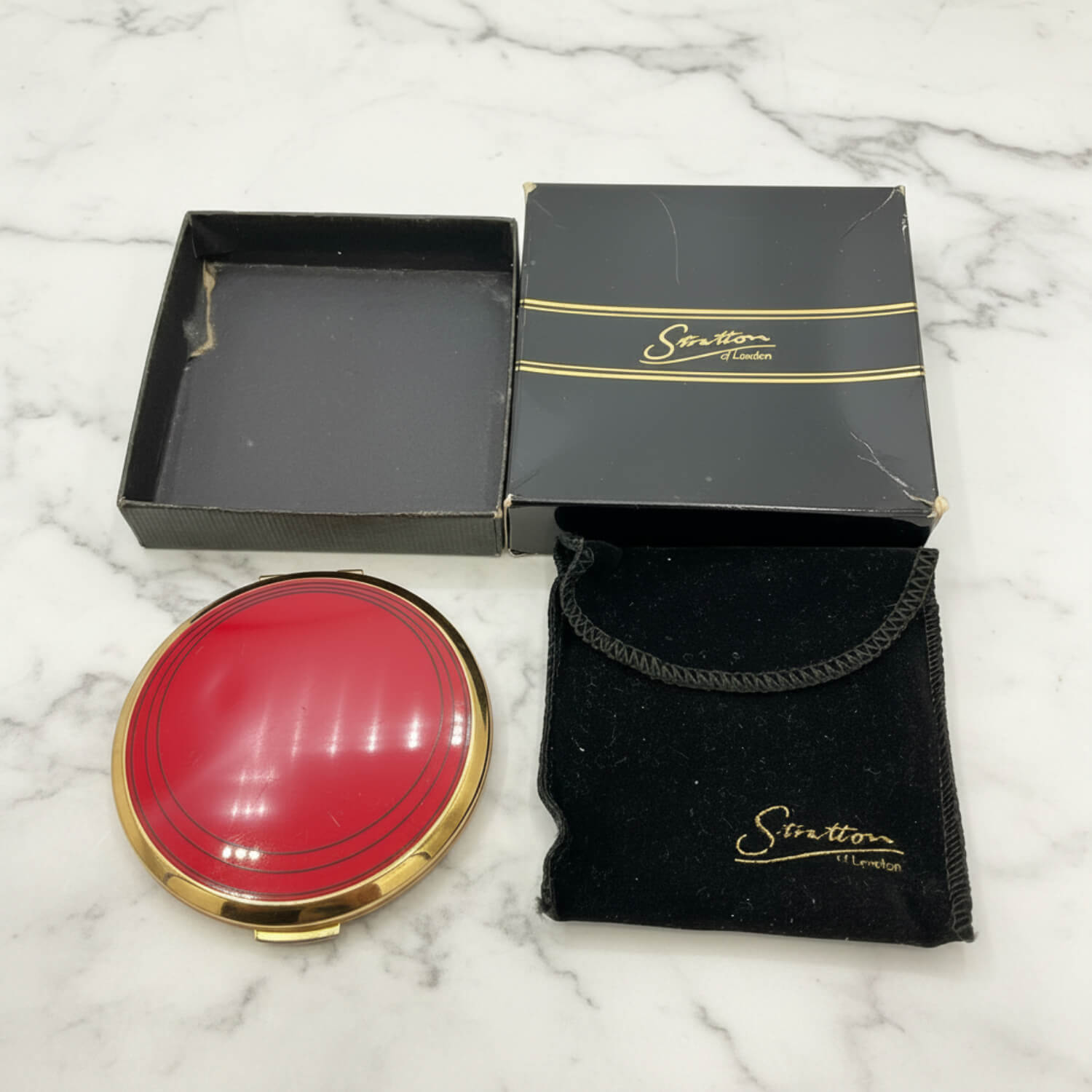 A round red compact mirror with a gold rim is displayed next to its black presentation box, inner box, and black velvet pouch with gold lettering, all on a white marble surface.