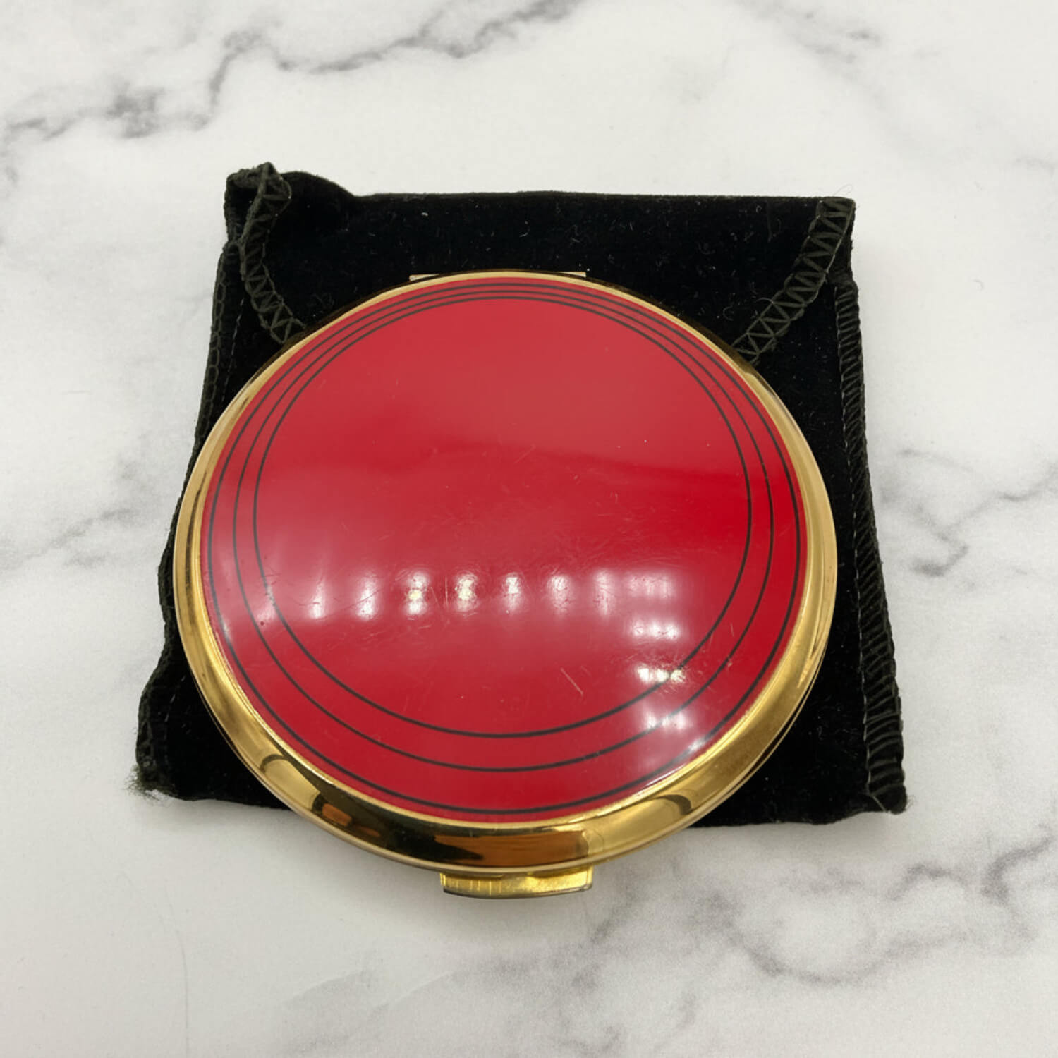 A round red compact mirror with a gold-tone metal rim, resting on a black velvet pouch over a white marble surface.
