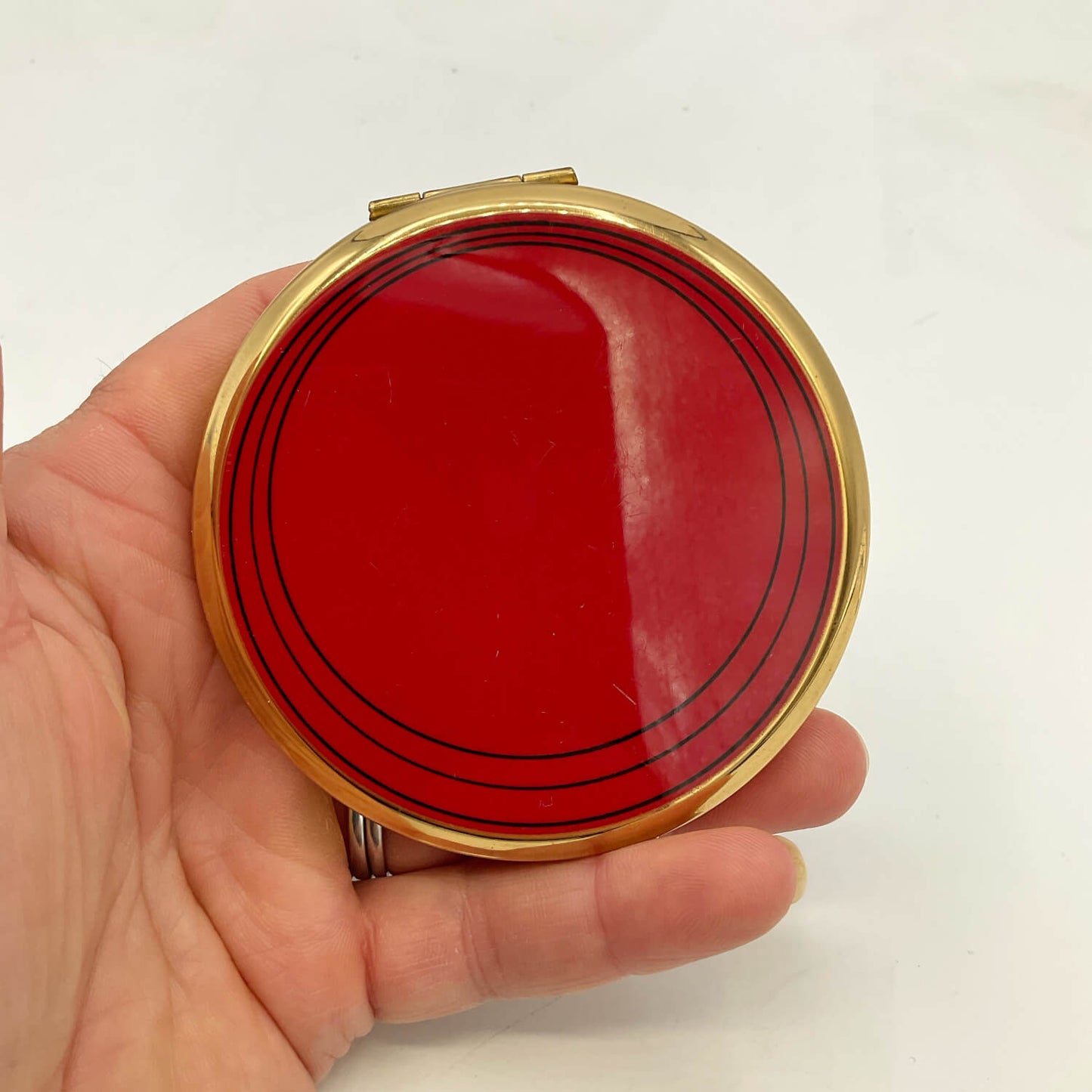 A round compact mirror with a gold-tone metal frame and a glossy red enamel face featuring black concentric circle lines, held in a person's hand against a plain white background.