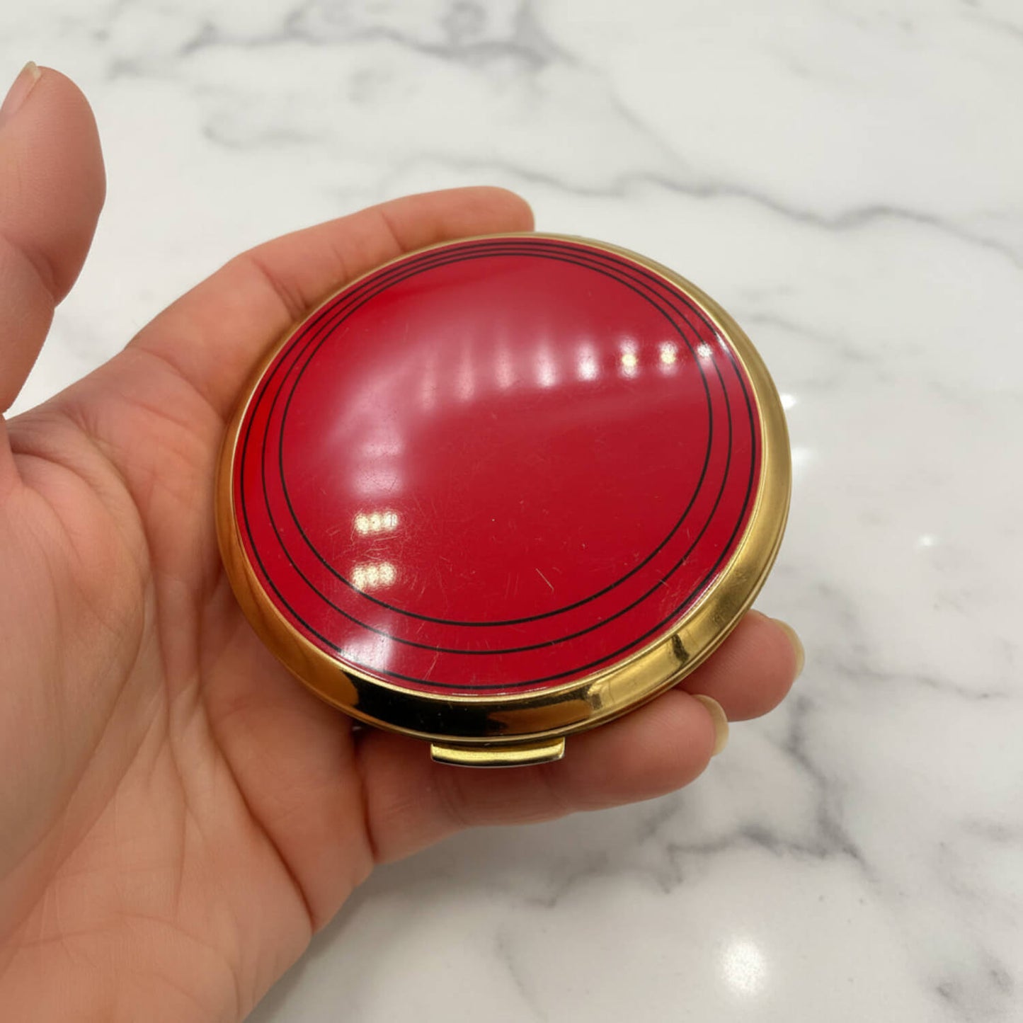A hand holding a round red compact case with a gold-tone metal rim, set against a white marble background.