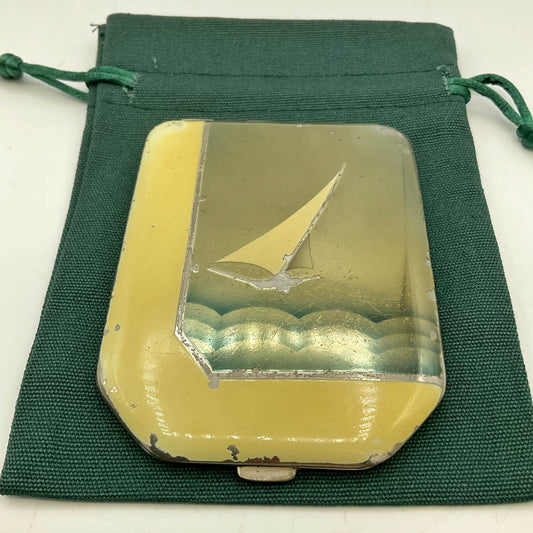 A vintage rectangular compact case with an Art Deco body and an etched sailboat design, displayed on a dark green drawstring pouch.
