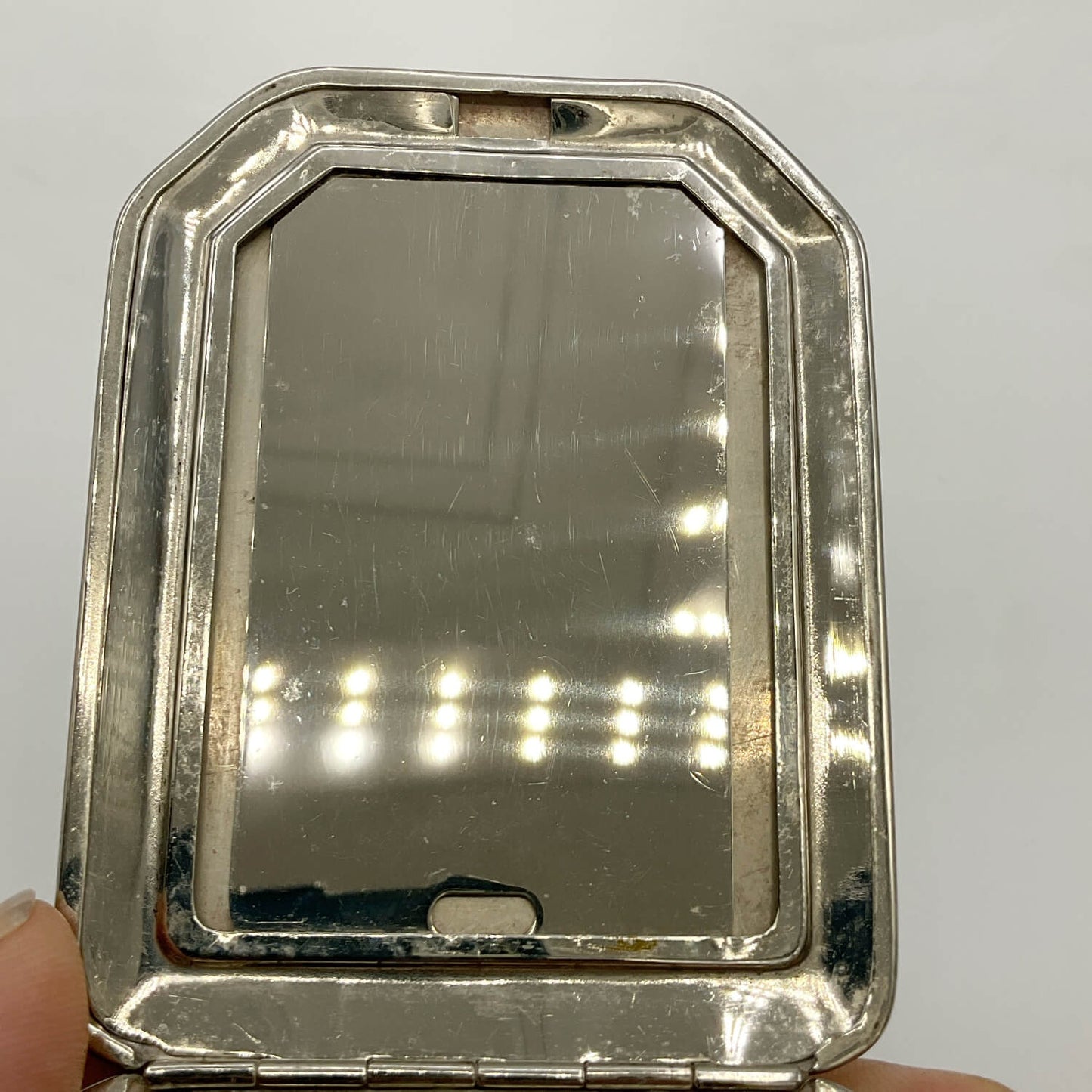 A vintage silver-toned compact mirror with a rounded rectangular frame, held open by fingers against a plain light background.