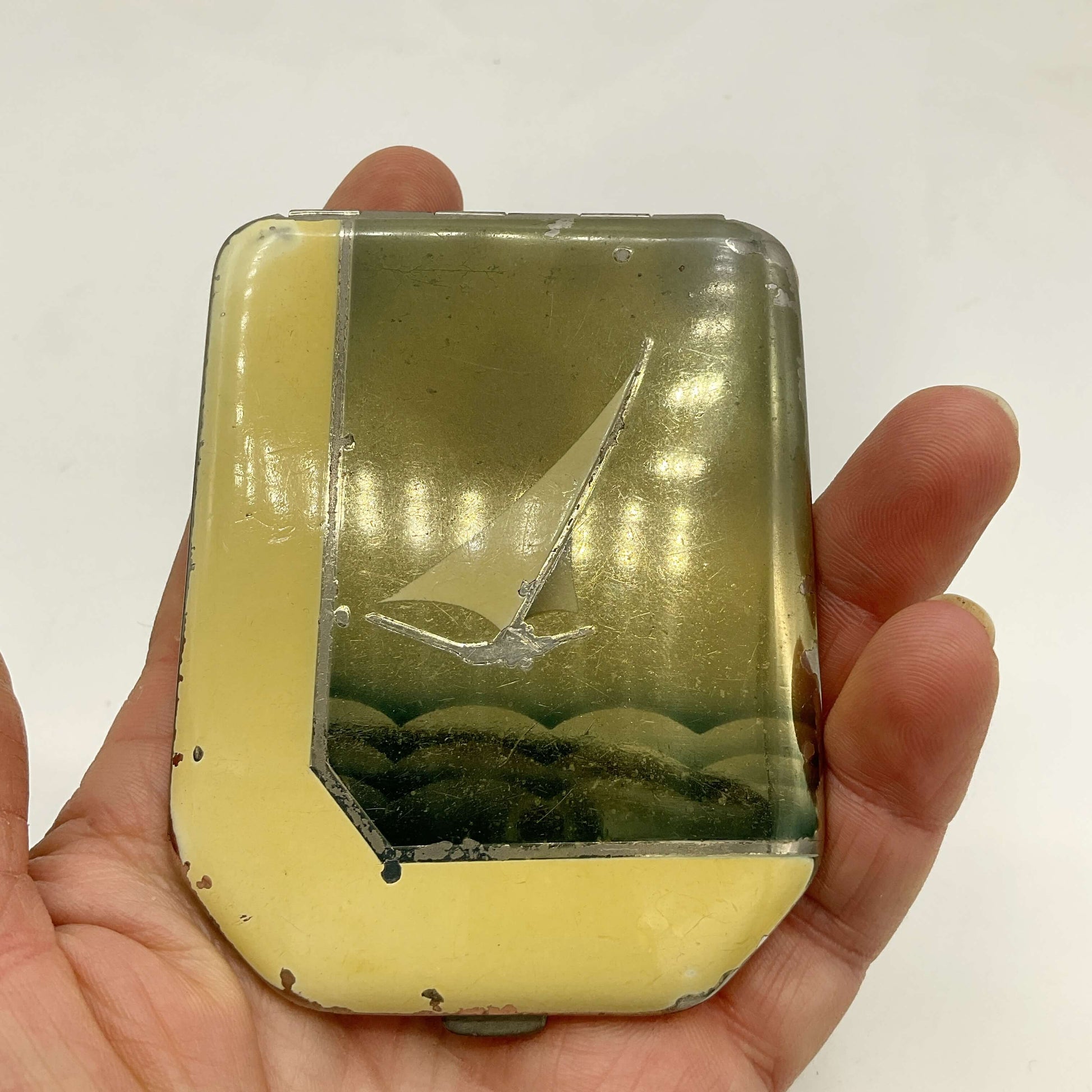 A vintage rectangular enamel compact held in a hand, featuring a yellow border and a central illustration of a sailing boat with a white background.