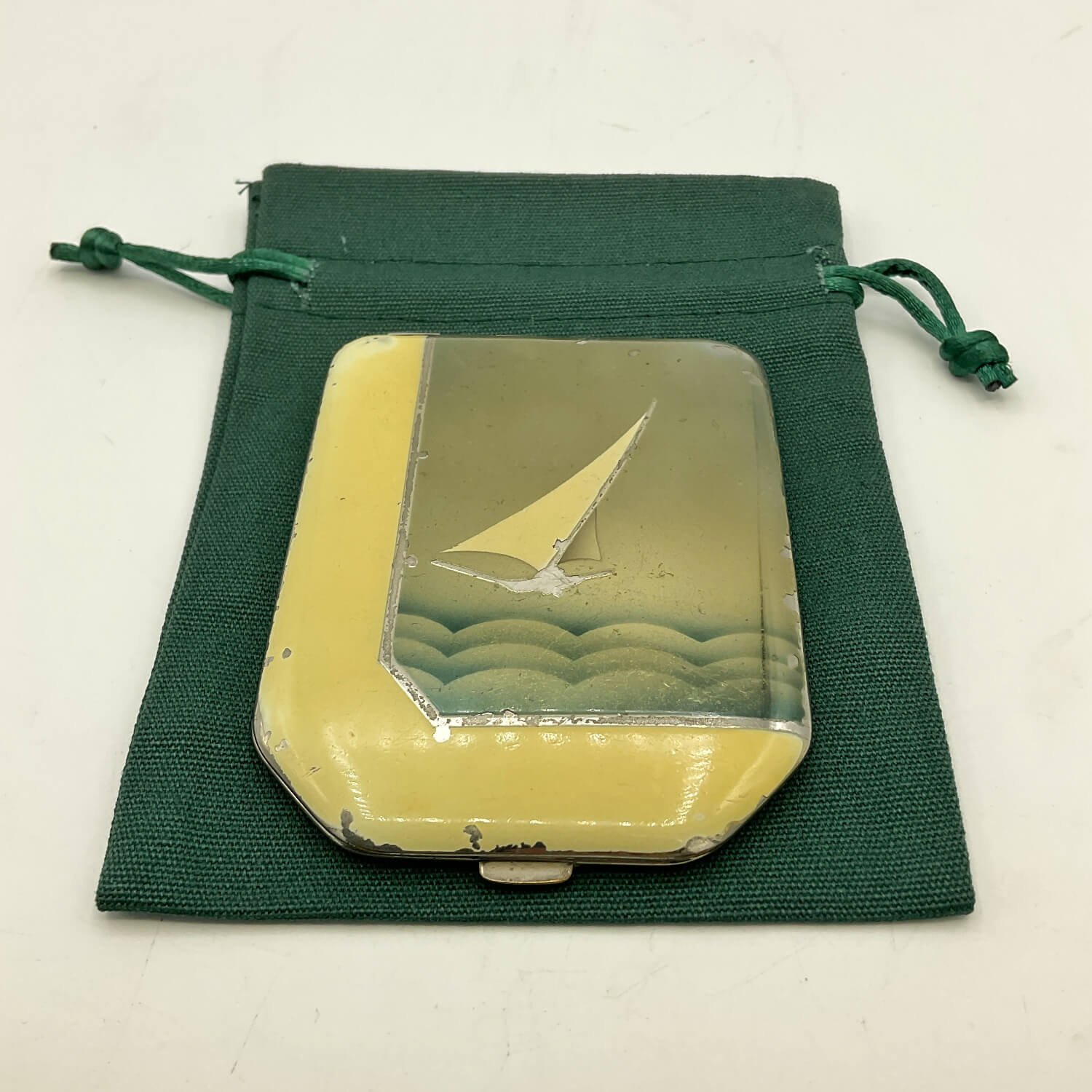 A vintage rectangular compact with a yellow enamel border and a clear top depicting a sailboat on waves, shown with a dark green drawstring pouch on a white surface.