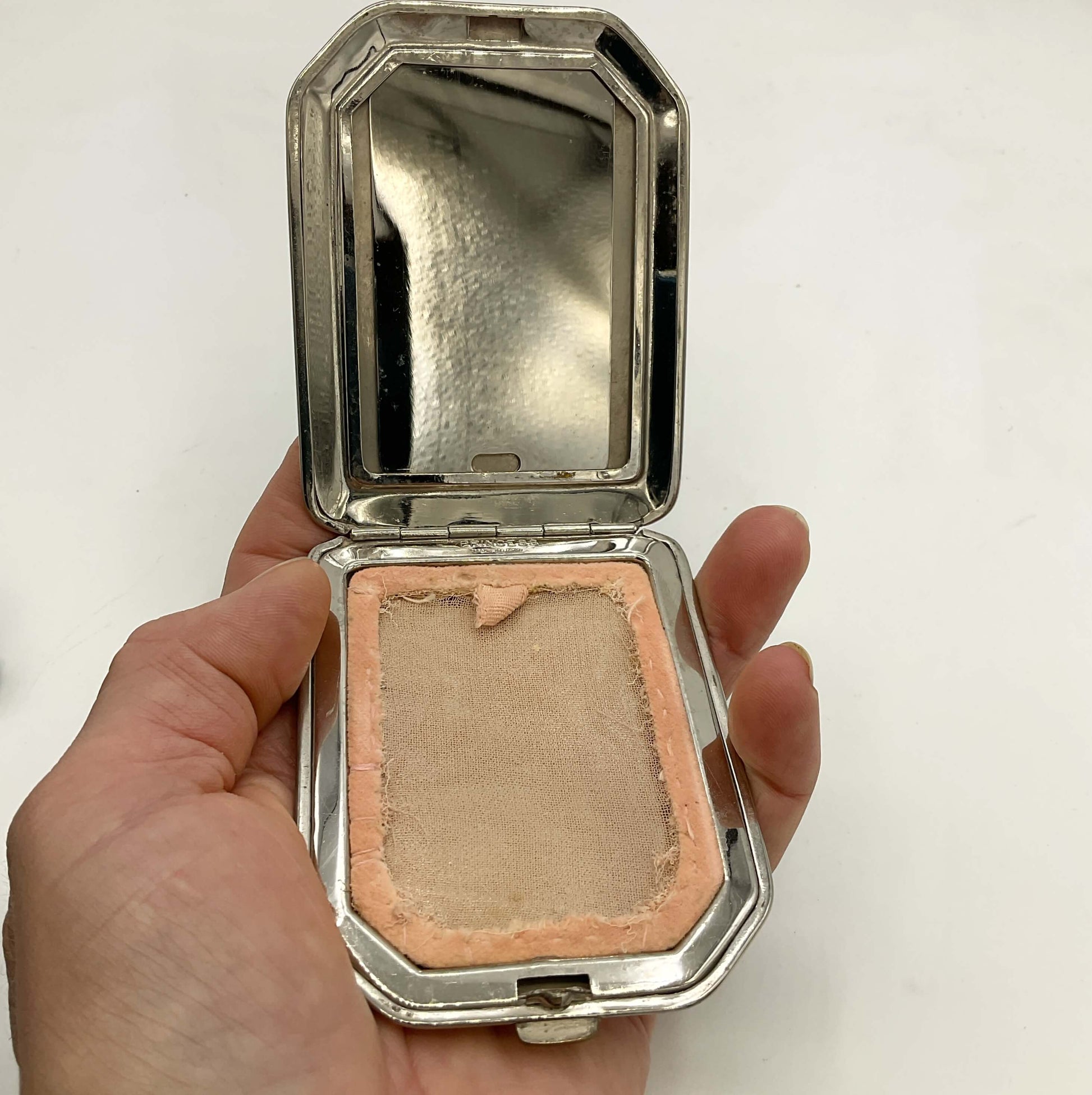An open vintage silver compact containing powder screen, held in a person's hand against a plain white background.