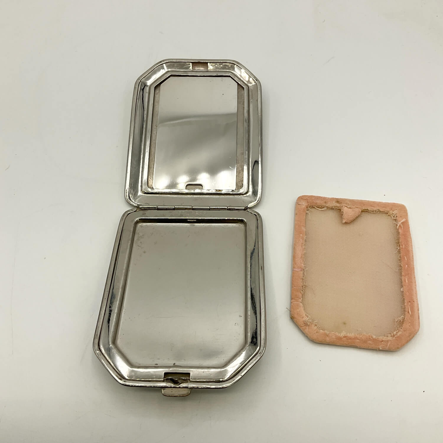 A vintage silver-toned metal compact case shown open, with its mirror lid facing up and a separate peach-colored fabric lining or insert placed beside it on a plain white background.