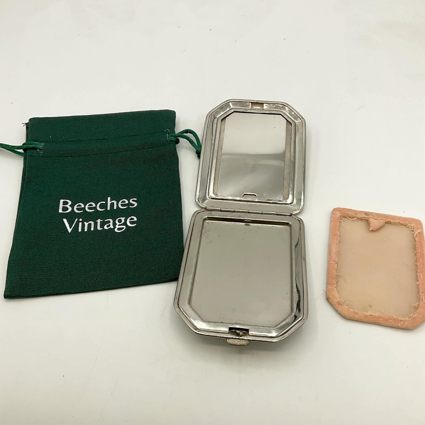 An open silver compact mirror with a peach-colored powder screen shown next to a dark green drawstring pouch printed with 'Beeches Vintage' on a white background.