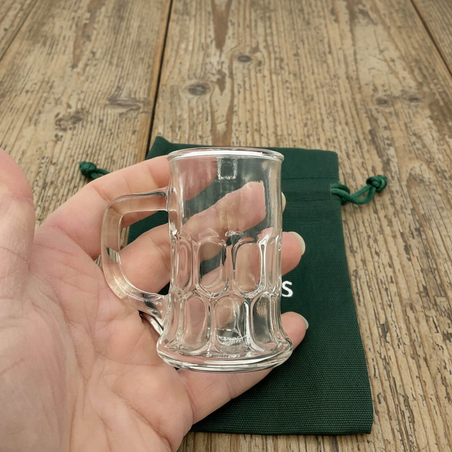 Clear glass mug held in a hand with a wooden surface and green drawstring bag in the background