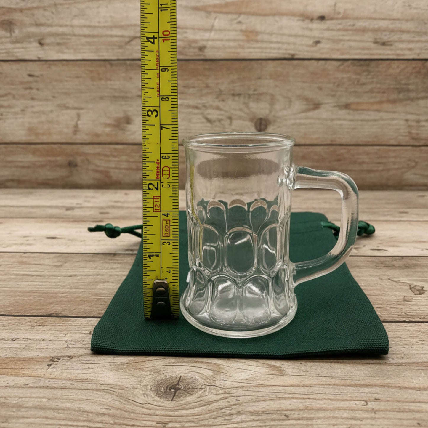 Clear glass mug with handle on a green coaster, measured against a ruler on a wooden surface.