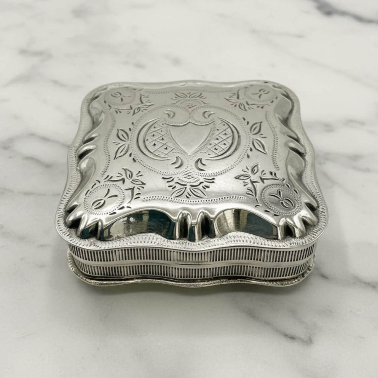 An ornate sterling silver jewelry box with a decorative lid featuring floral engravings and a central crest, resting on a white marble surface.