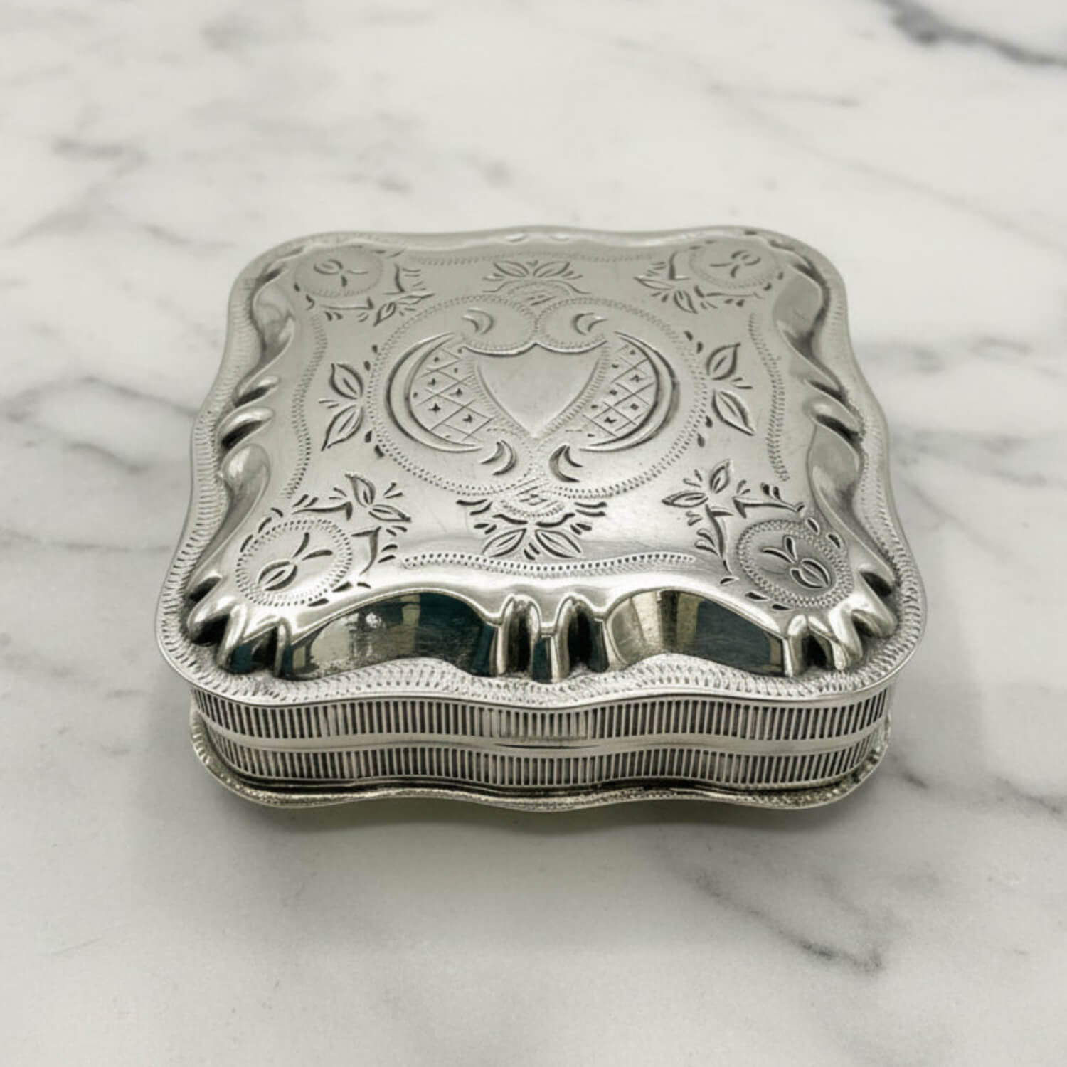 An ornate sterling silver jewelry box with a decorative lid featuring floral engravings and a central crest, resting on a white marble surface.