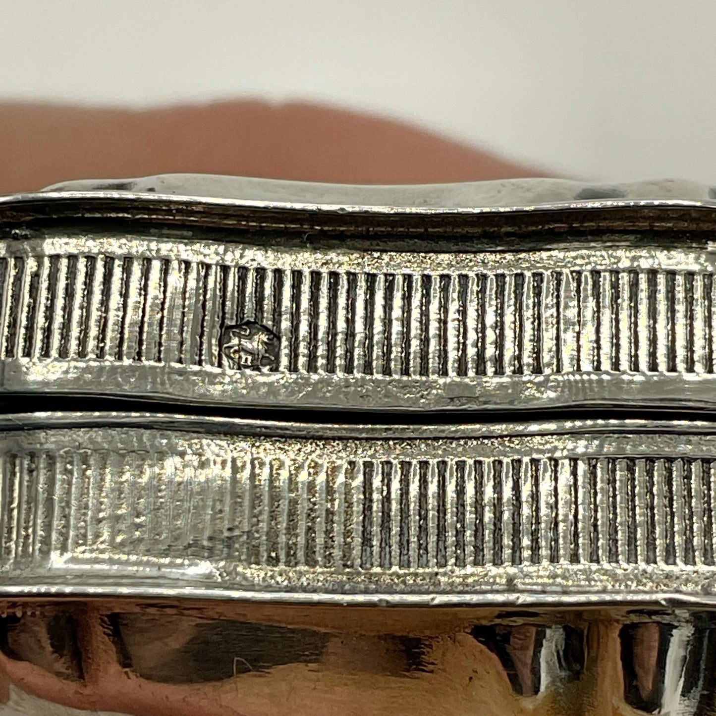 Close-up of a silver pill box with a ribbed textured surface and a small stamped hallmark shown against a soft blurred background.