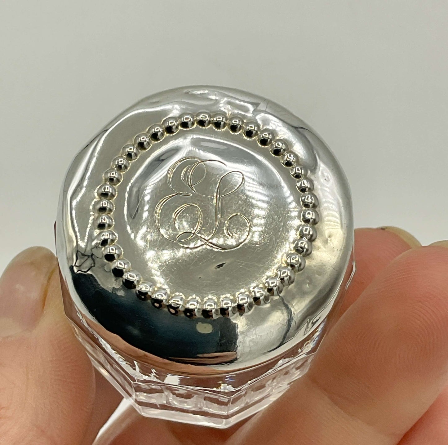 Silver hexagonal container with 'EL' monogram held between fingers against a light background