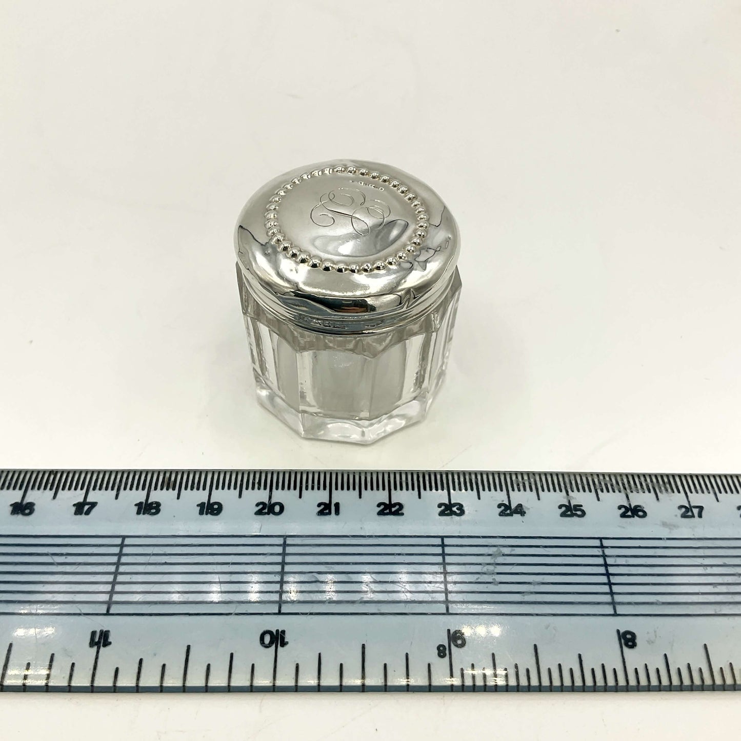 Small glass jar with a silver lid on a white background with a ruler for scale.