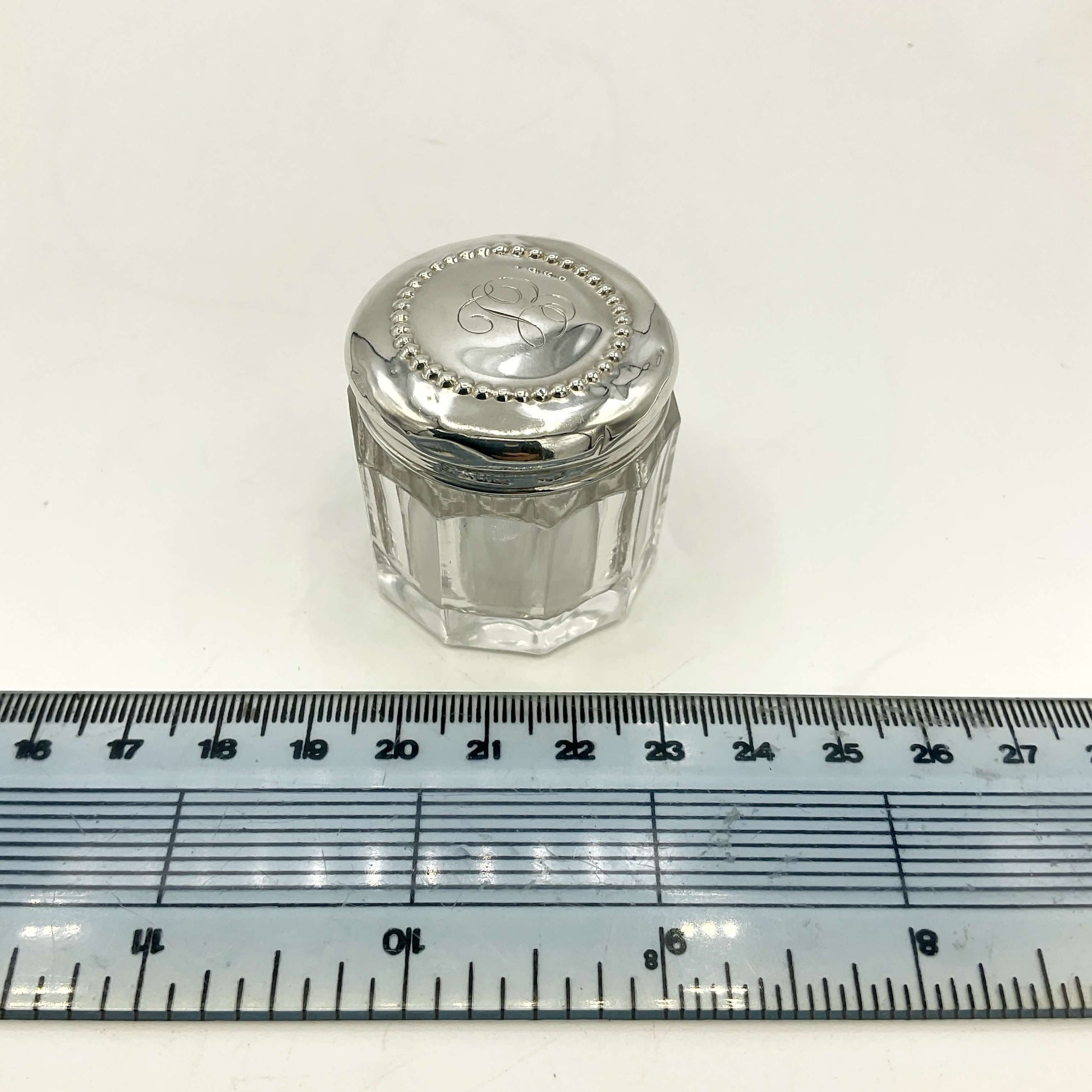 Small glass jar with a silver lid on a white background with a ruler for scale.