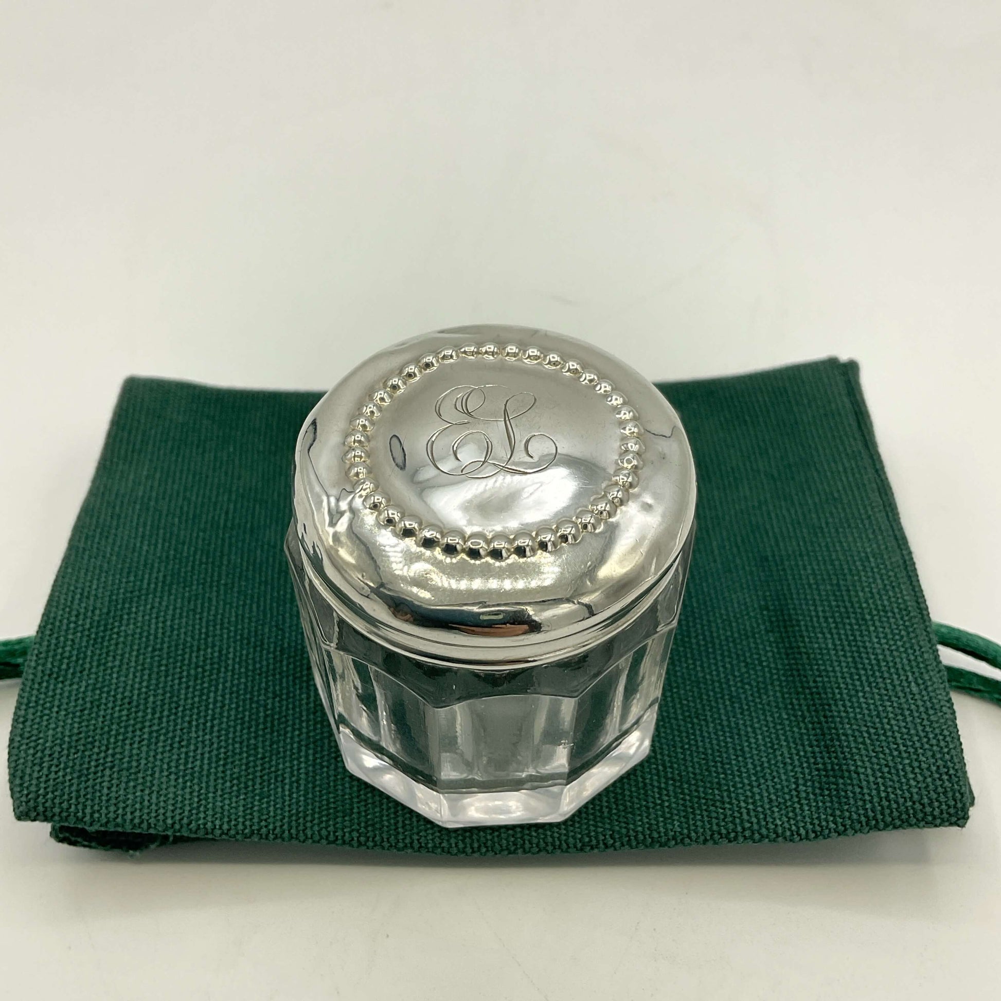 Silver lid with 'EL' initials on a glass container, placed on a green cloth.
