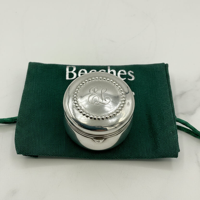 Silver lidded jar with monogram on a green fabric pouch labeled 'Beeches'.