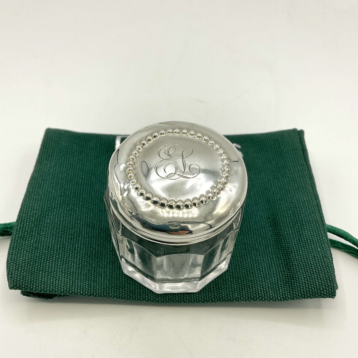 Silver jar with 'EL' engraving on a green cloth against a white background