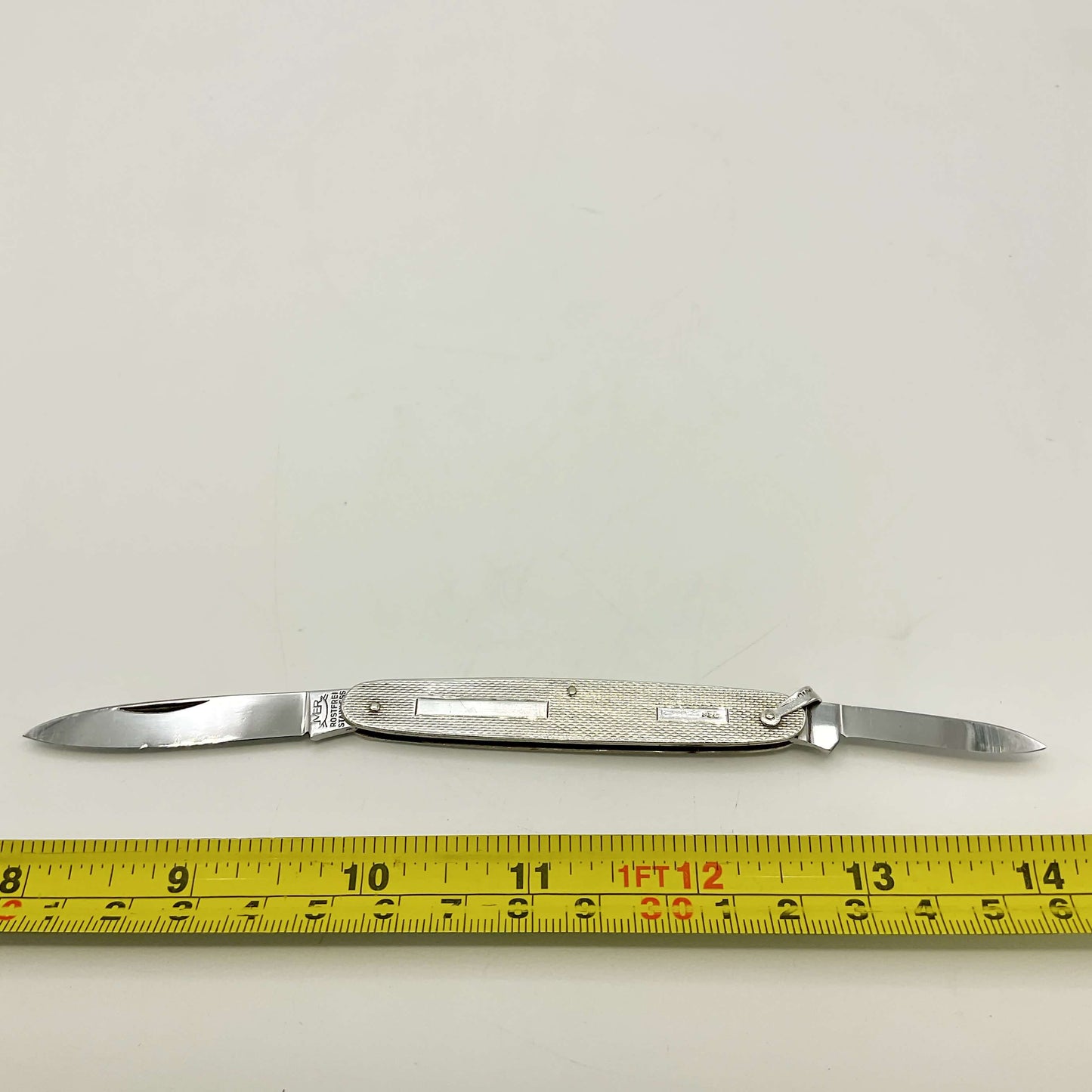Pen knife with two blades on a white background, measured by a yellow tape measure.