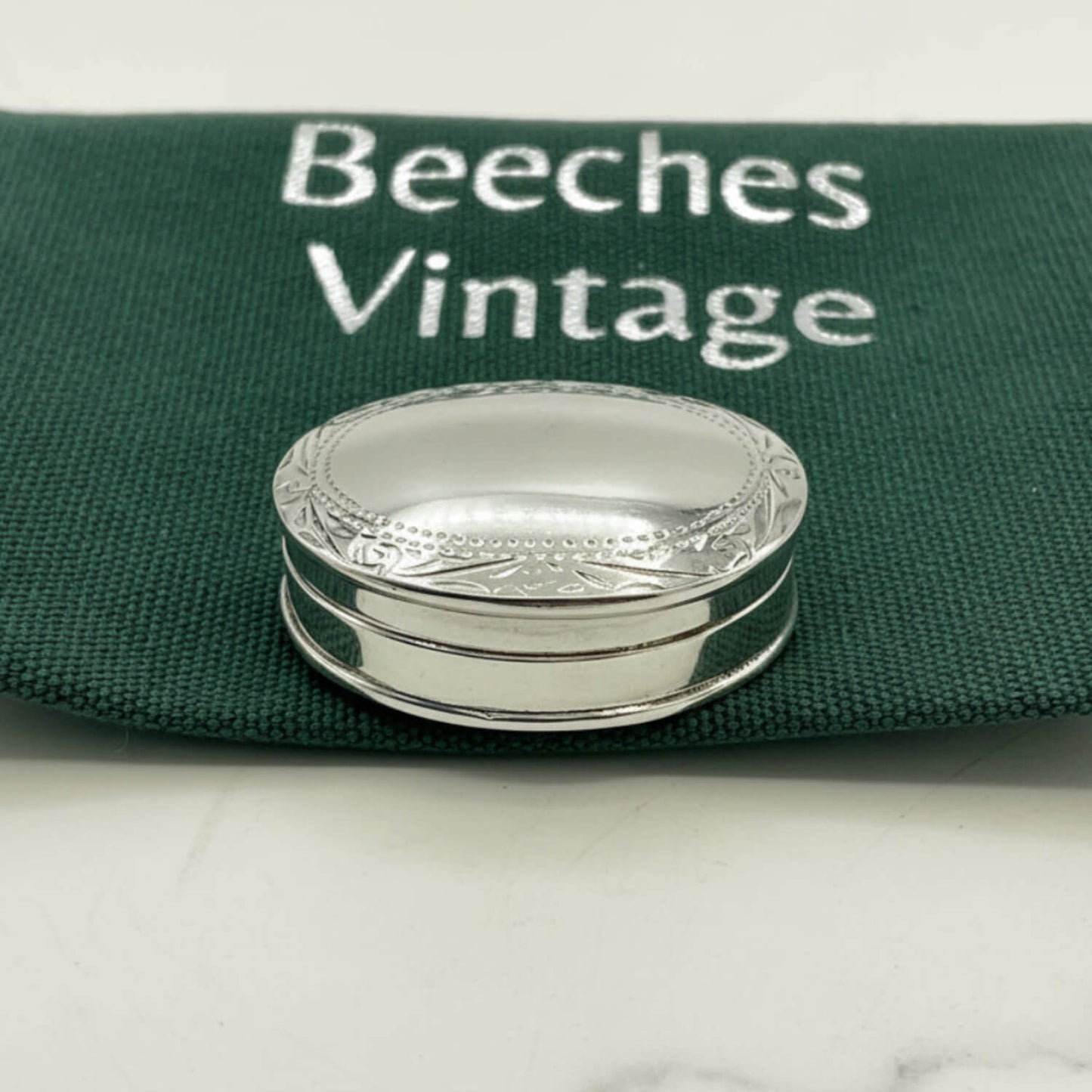 A small oval silver pill box with engraved detailing, resting on a dark green fabric pouch printed with the words 'Beeches Vintage'.