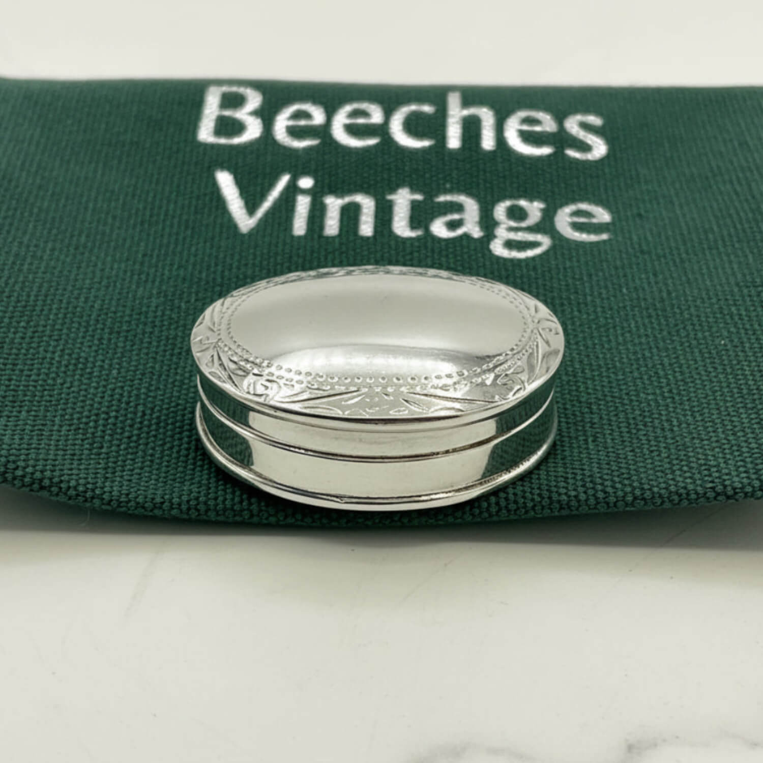 A small oval silver pill box with engraved detailing, resting on a dark green fabric pouch printed with the words 'Beeches Vintage'.