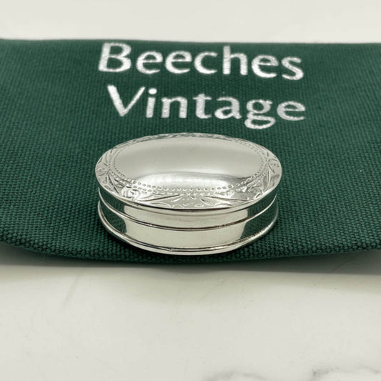 A small oval silver pill box with engraved detailing, resting on a dark green fabric pouch printed with the words 'Beeches Vintage'.