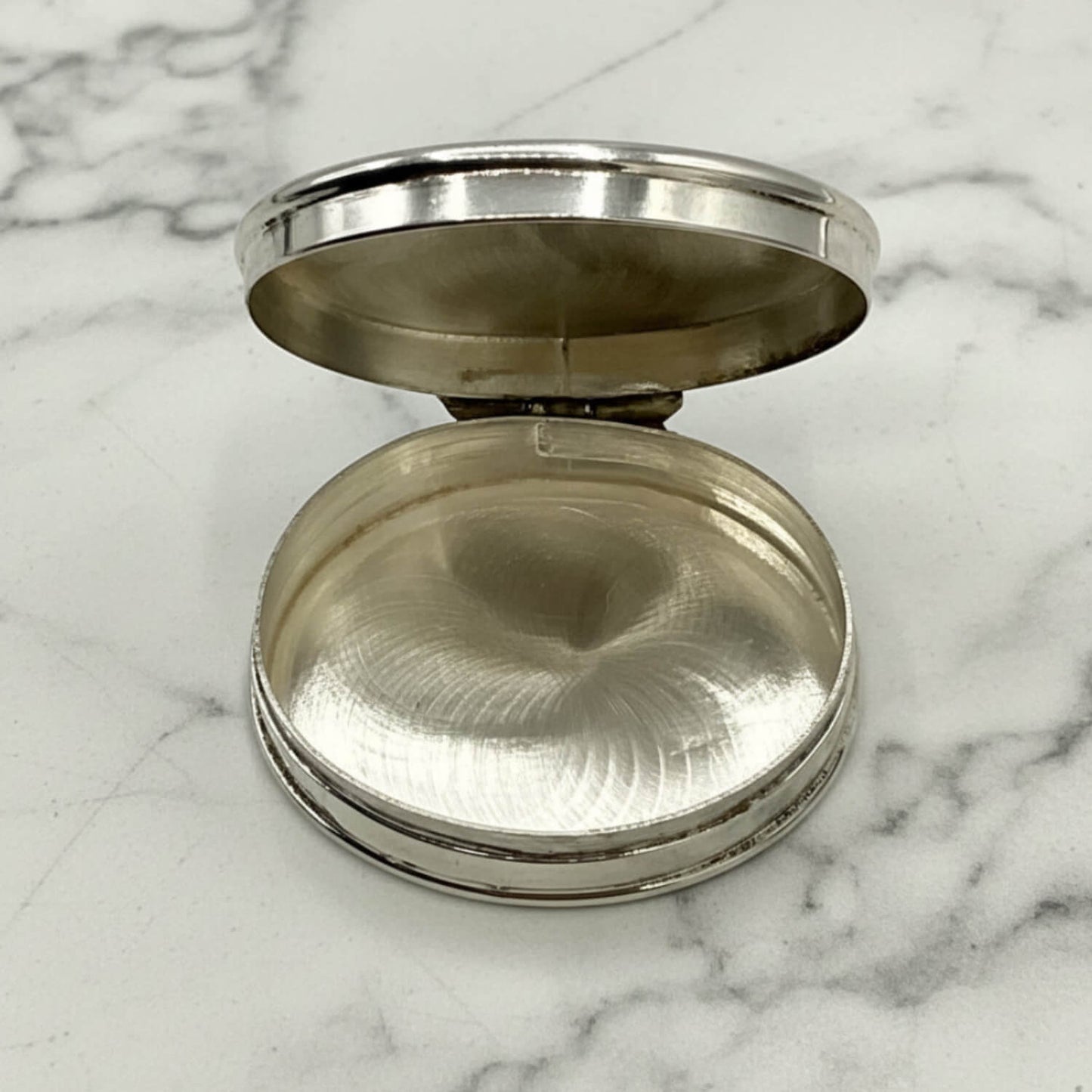 A oval silver pill case, shown open on a white marble surface. The case has a brushed metallic finish and a polished rim.