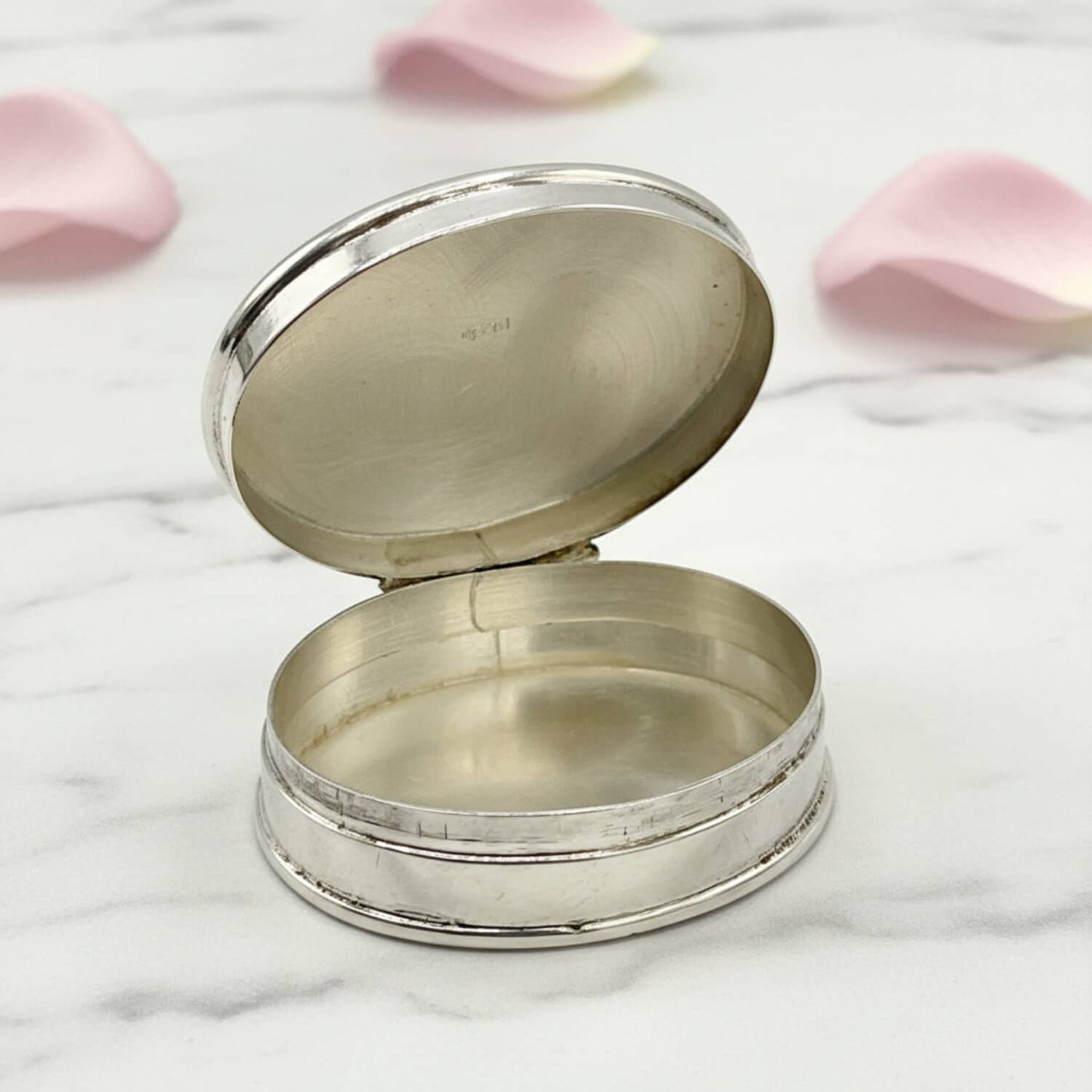 An open oval silver pill box with a hinged lid, photographed on a white marble surface with soft pink petals in the background.