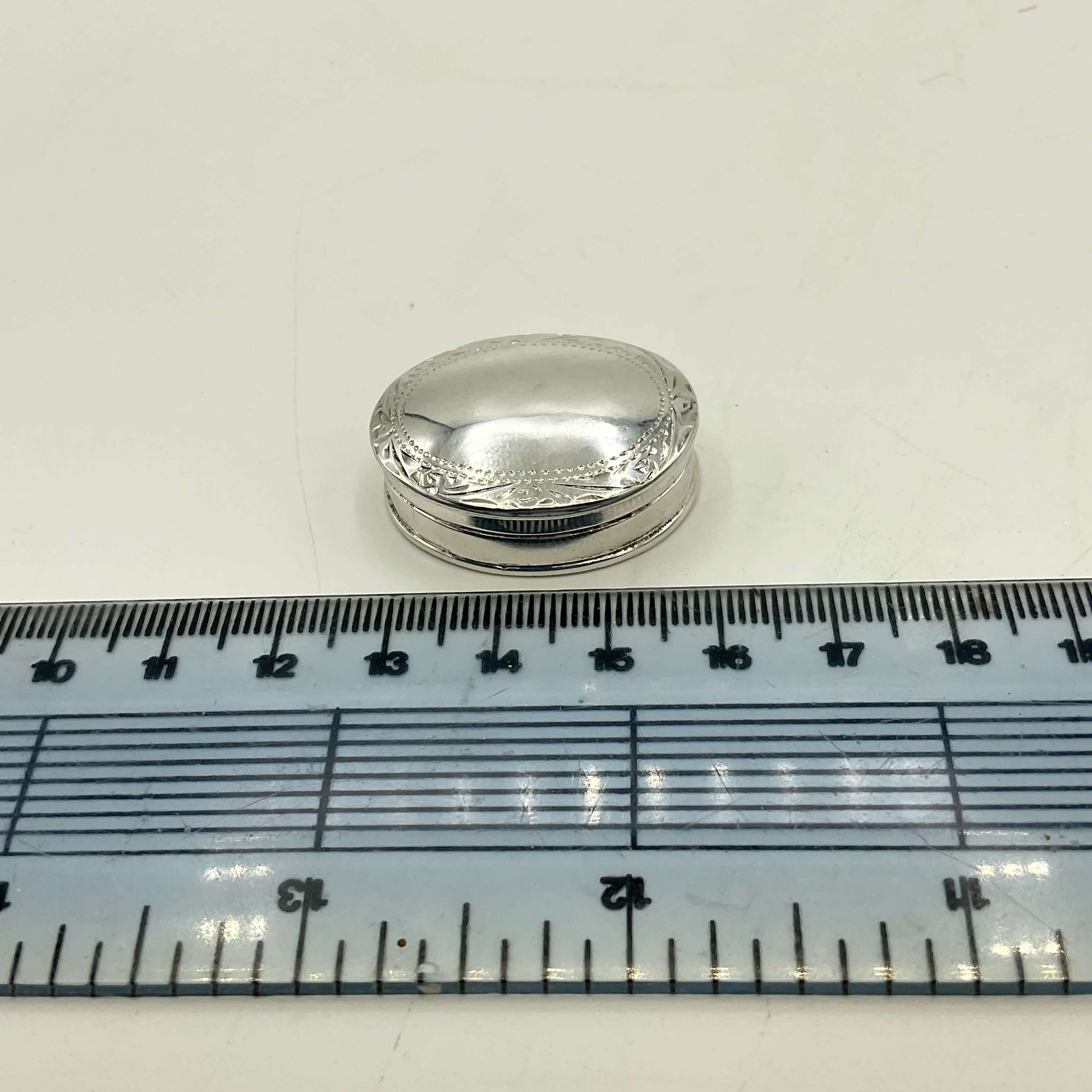 A small oval silver pill box with a domed lid sits above a blue measuring ruler on a white surface, showing its size.