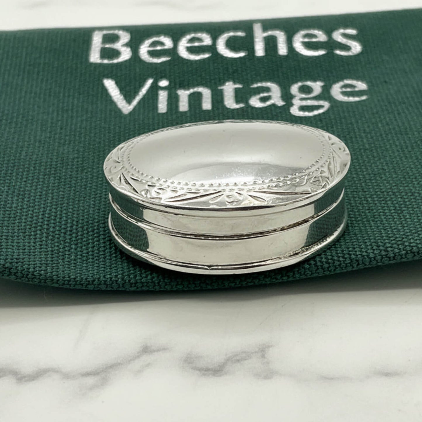 A small oval silver pill box with engraved decorative detailing around the rim, placed on a dark green fabric surface printed with the words 'Beeches Vintage'.