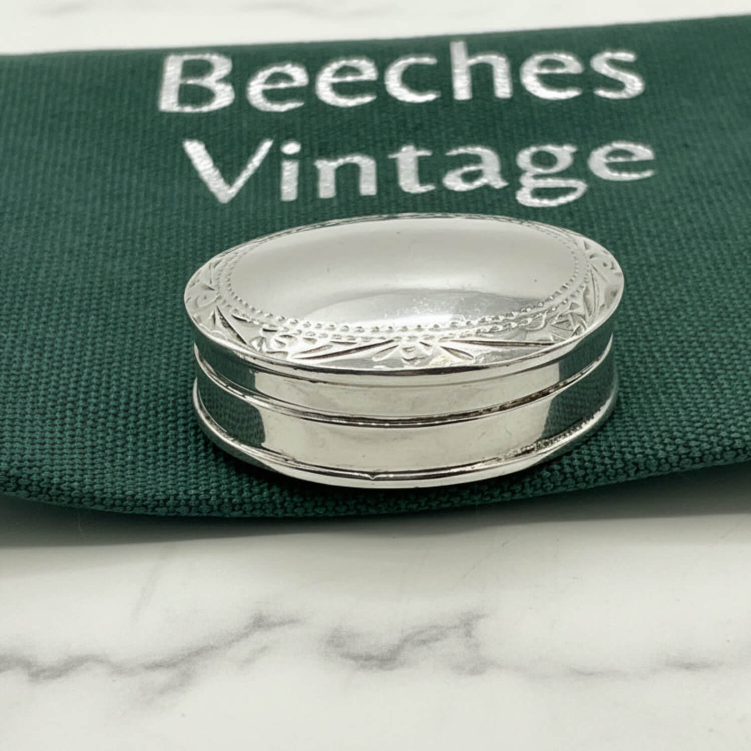 A small oval silver pill box with engraved decorative detailing around the rim, placed on a dark green fabric surface printed with the words 'Beeches Vintage'.