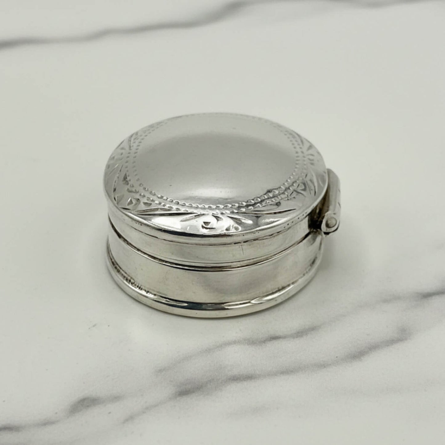 A small round silver pill box with a domed lid and engraved decorative border, photographed on a light marble-like surface.
