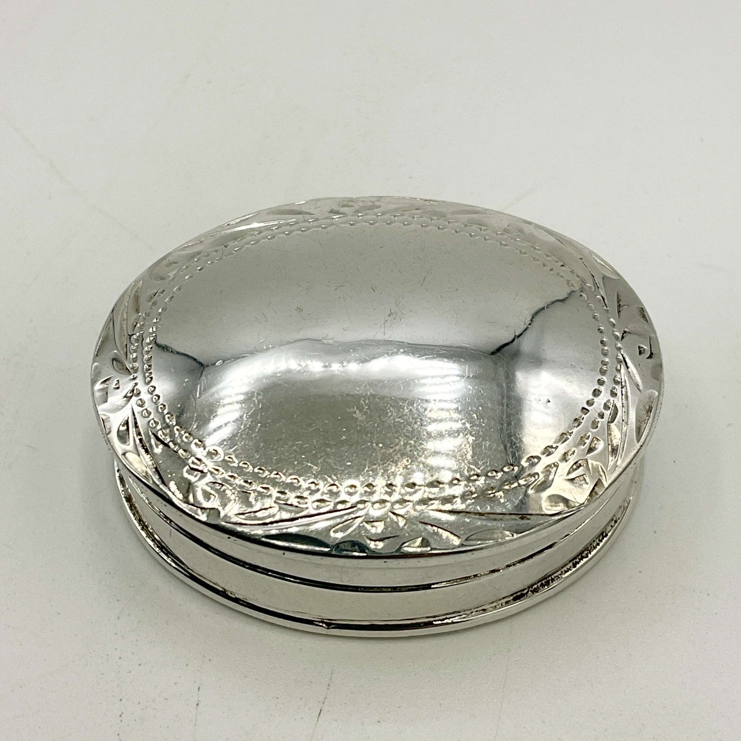 A small oval silver pill box with a hinged lid, featuring decorative embossed floral and beaded border details around the rim, shown on a plain light background.