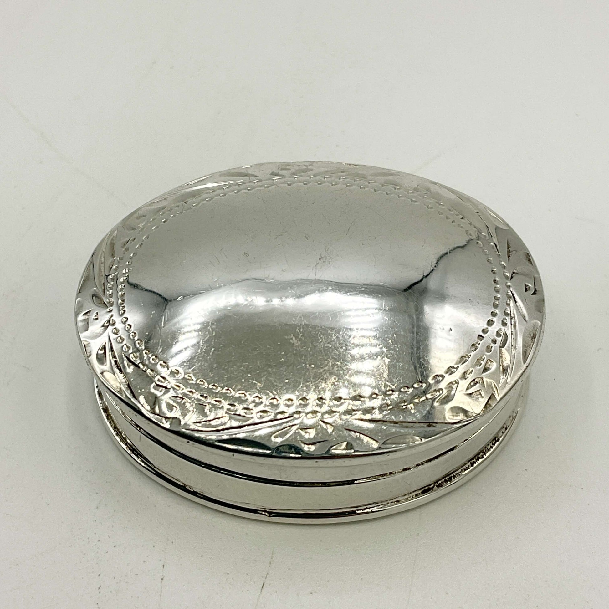 A small oval silver pill box with a hinged lid, featuring decorative embossed floral and beaded border details around the rim, shown on a plain light background.
