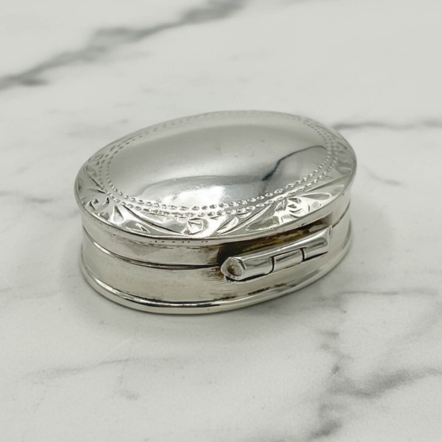 A small oval silver pill box with engraved decorative detailing on the lid, resting on a white marble surface.