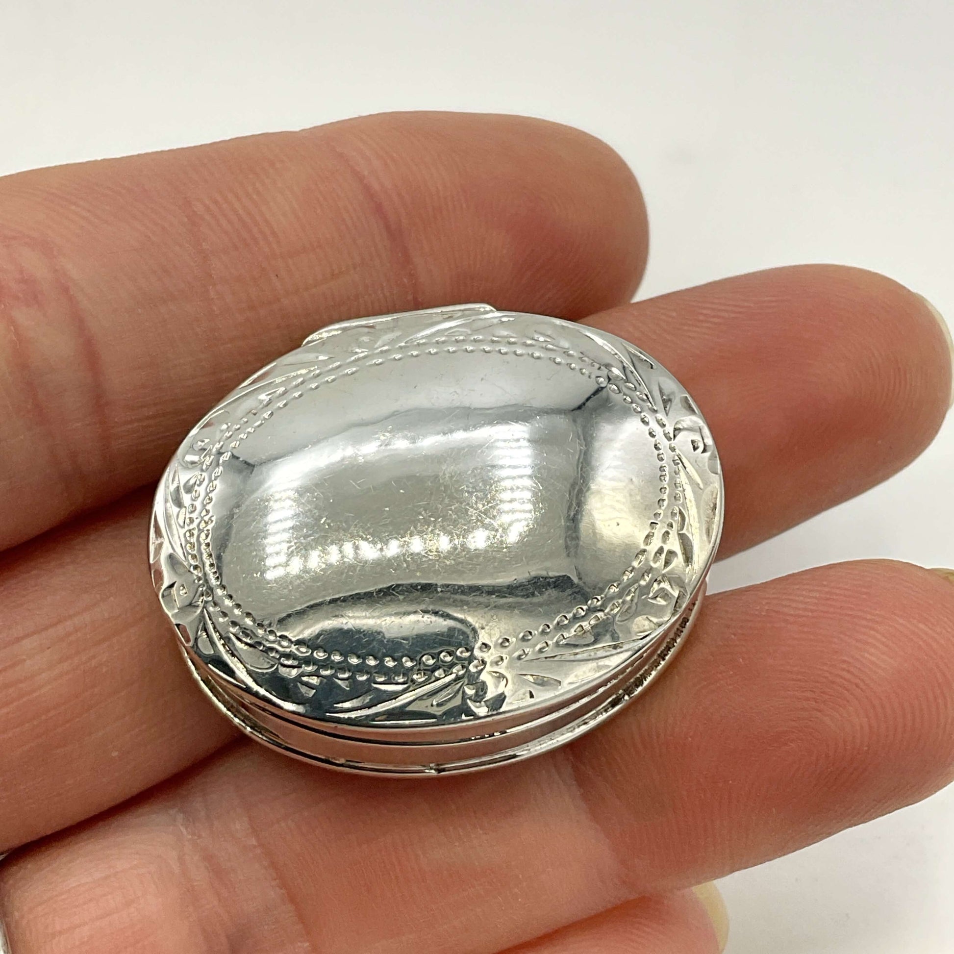 A small oval silver pill case with engraved decorative detailing, held in a person's fingers against a plain light background.