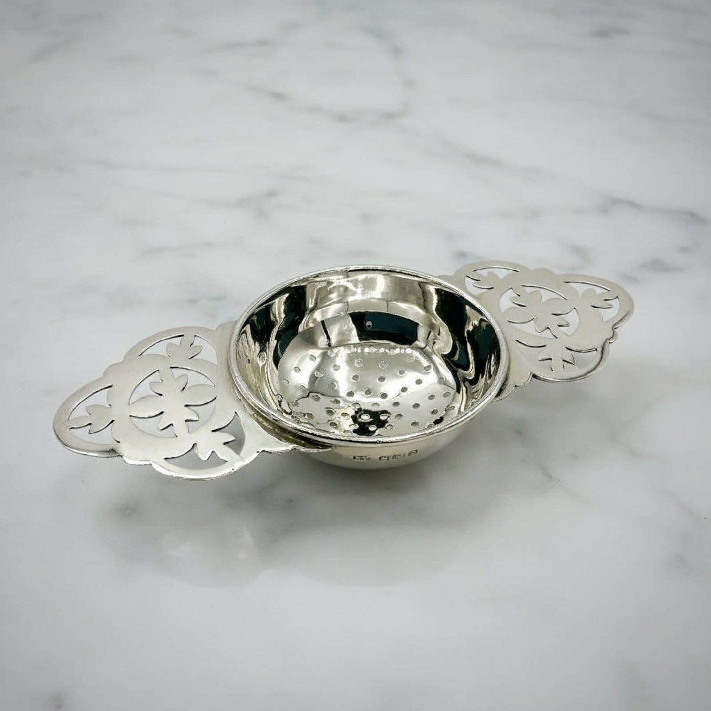 Silver decorative tea strainer with intricate designs on a marble surface