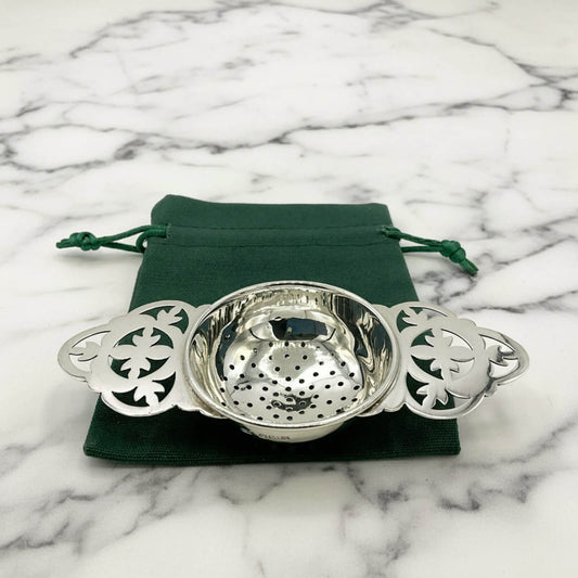 Silver tea strainer with decorative patterns on a green drawstring bag against a marble background
