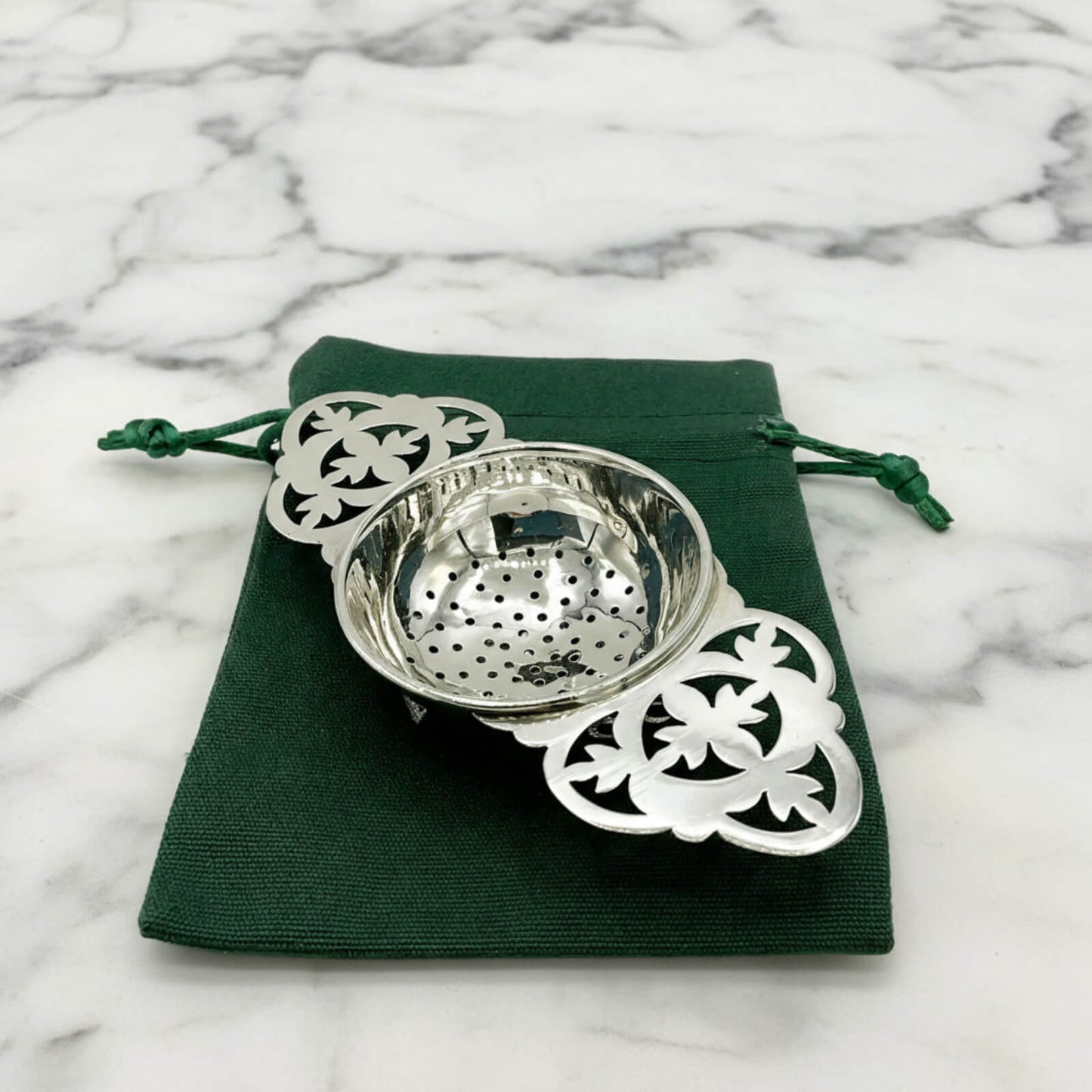 Silver tea strainer with decorative elements on a green cloth against a marble background