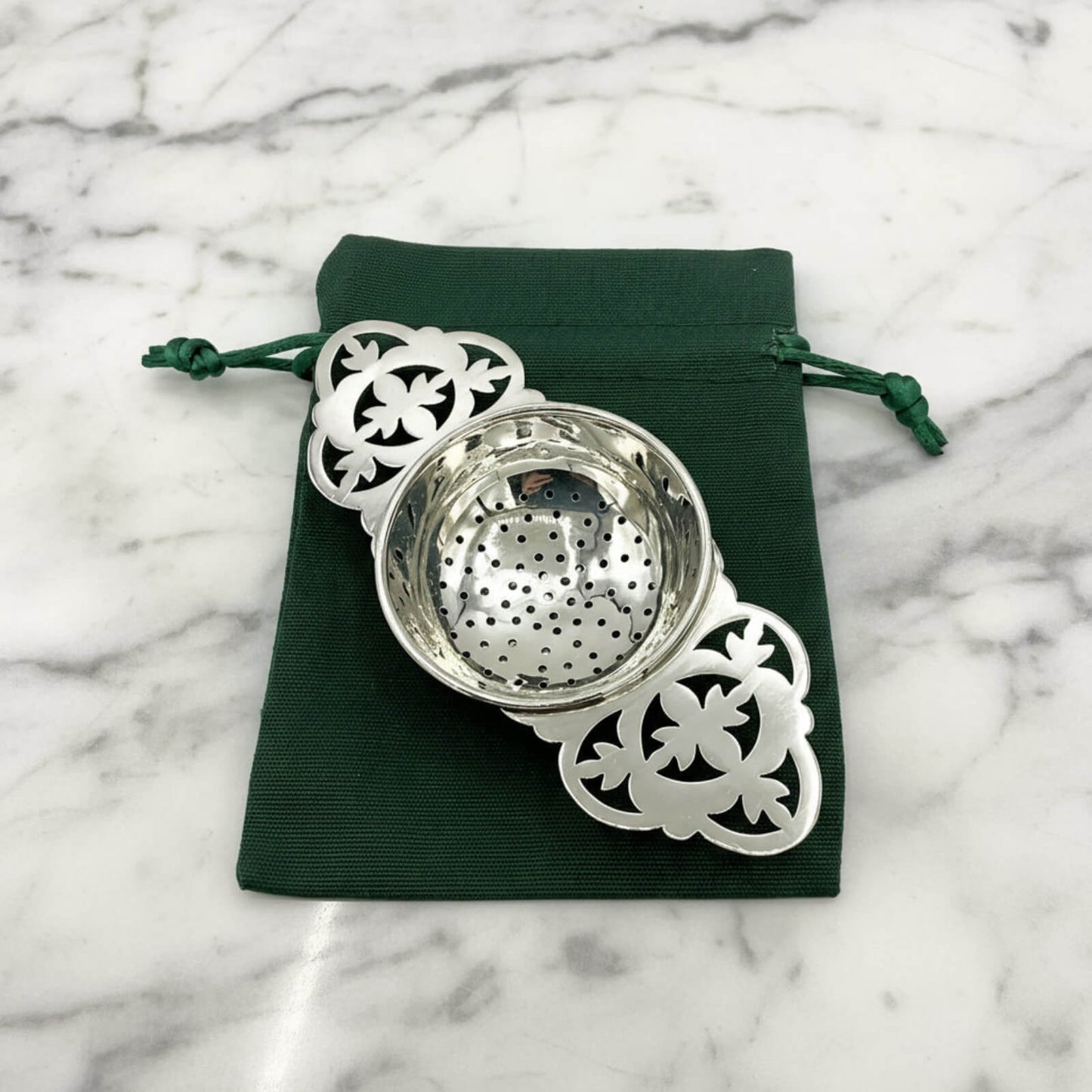 Silver tea strainer with decorative elements on a green drawstring bag against a marble background