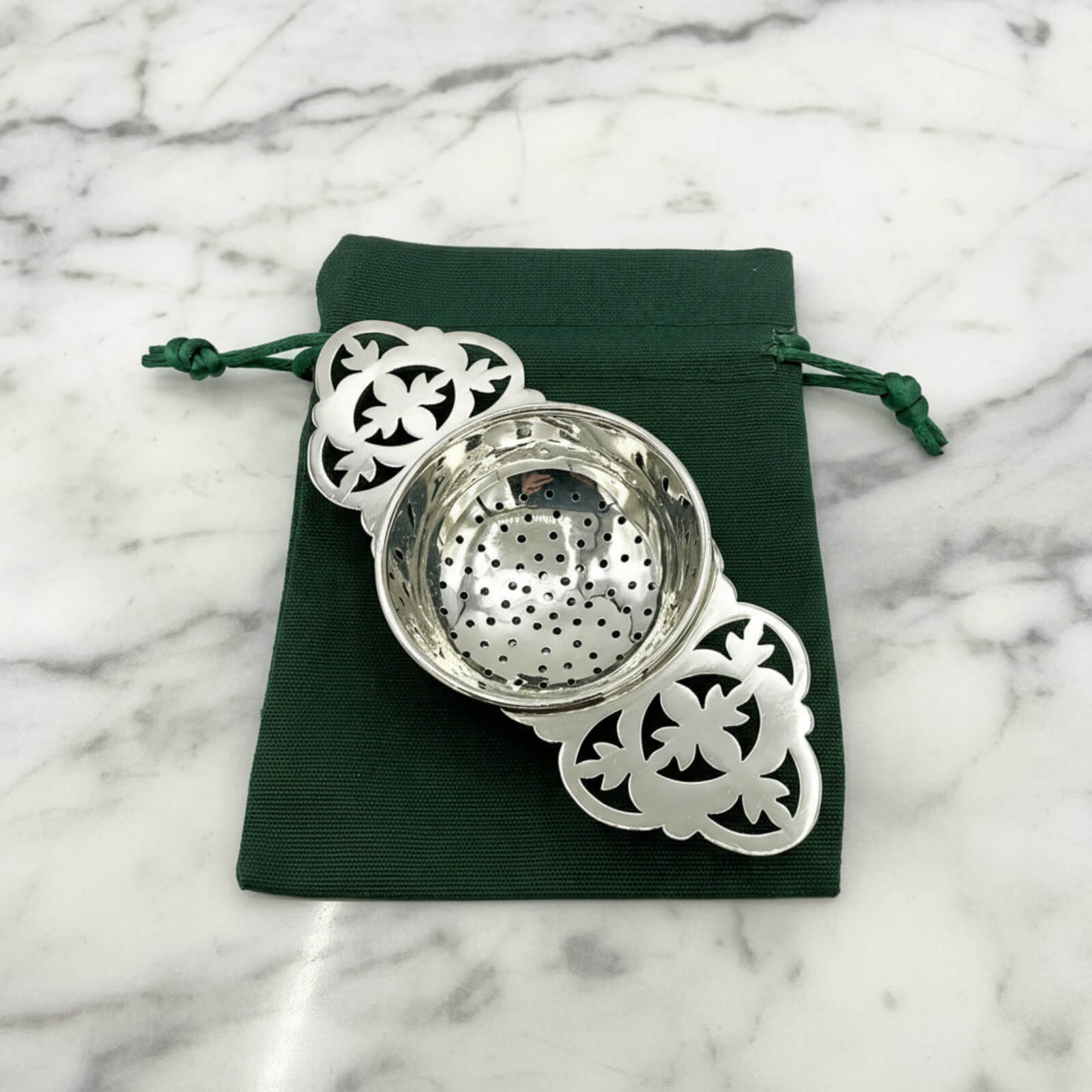 Silver tea strainer with decorative elements on a green drawstring bag against a marble background