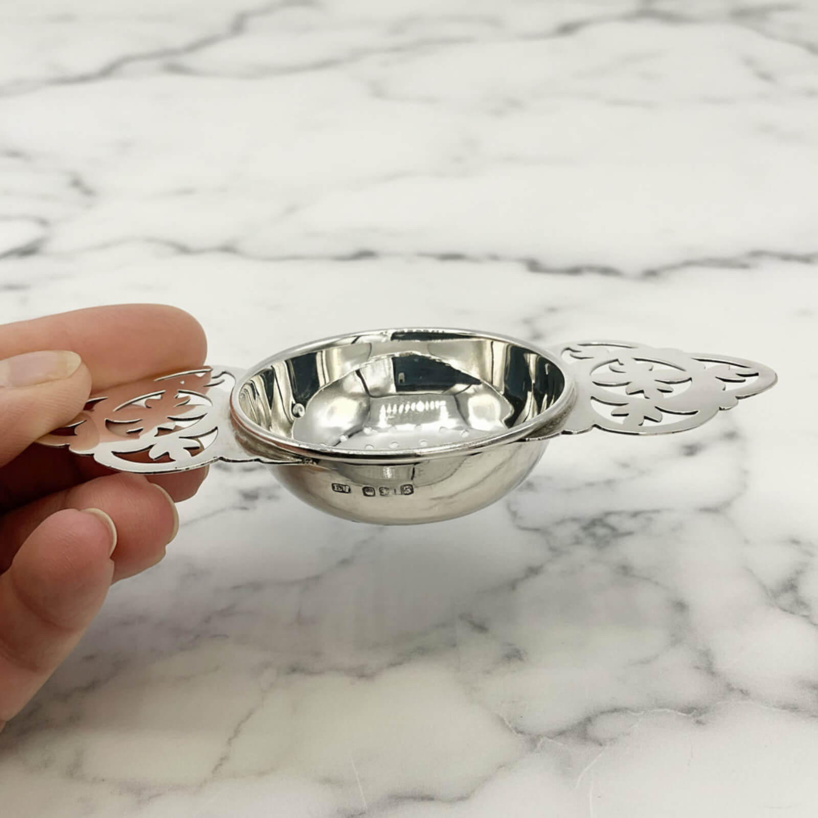 Silver tea strainer held by a hand on a marble background