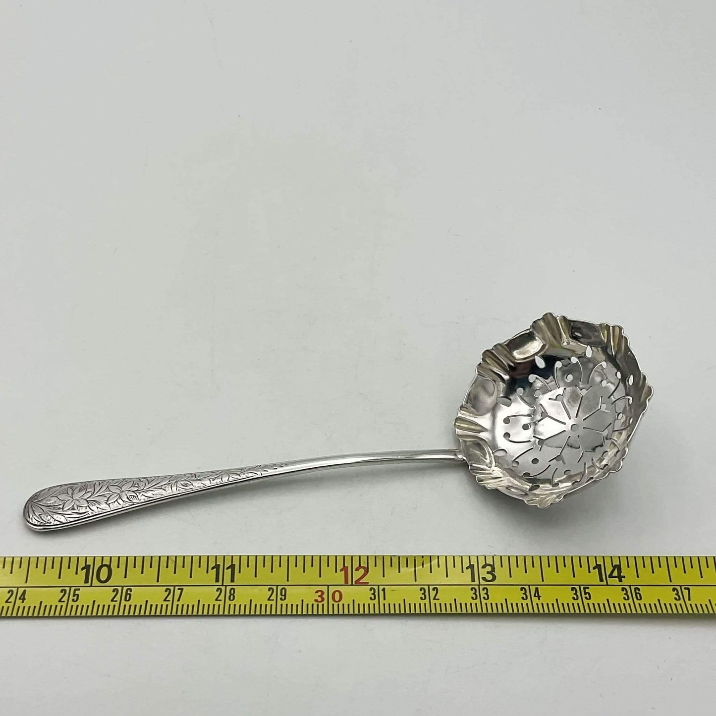 Silver sugar sifter with decorative handle on a white background, measured against a ruler.