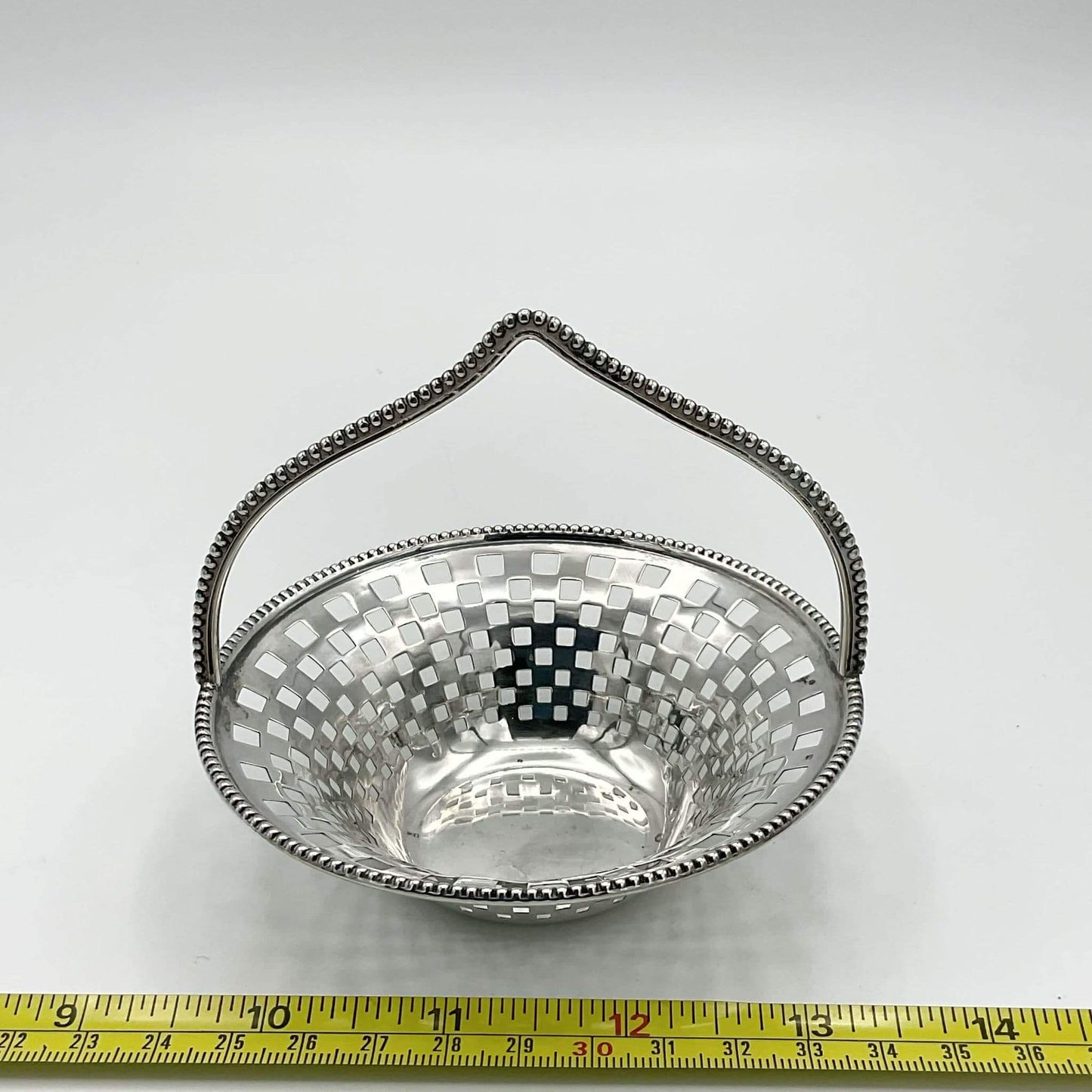 Silver basket with a handle on a white background, measured against a tape measure.