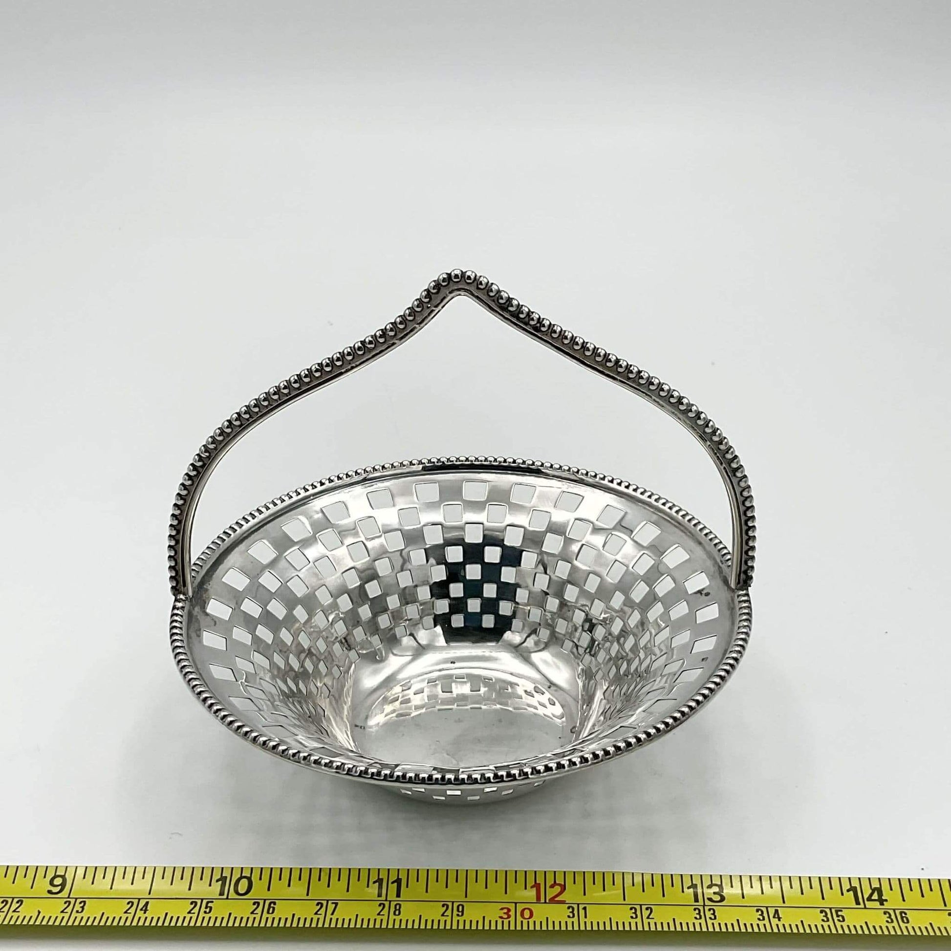 Silver basket with a handle on a white background, measured against a tape measure.