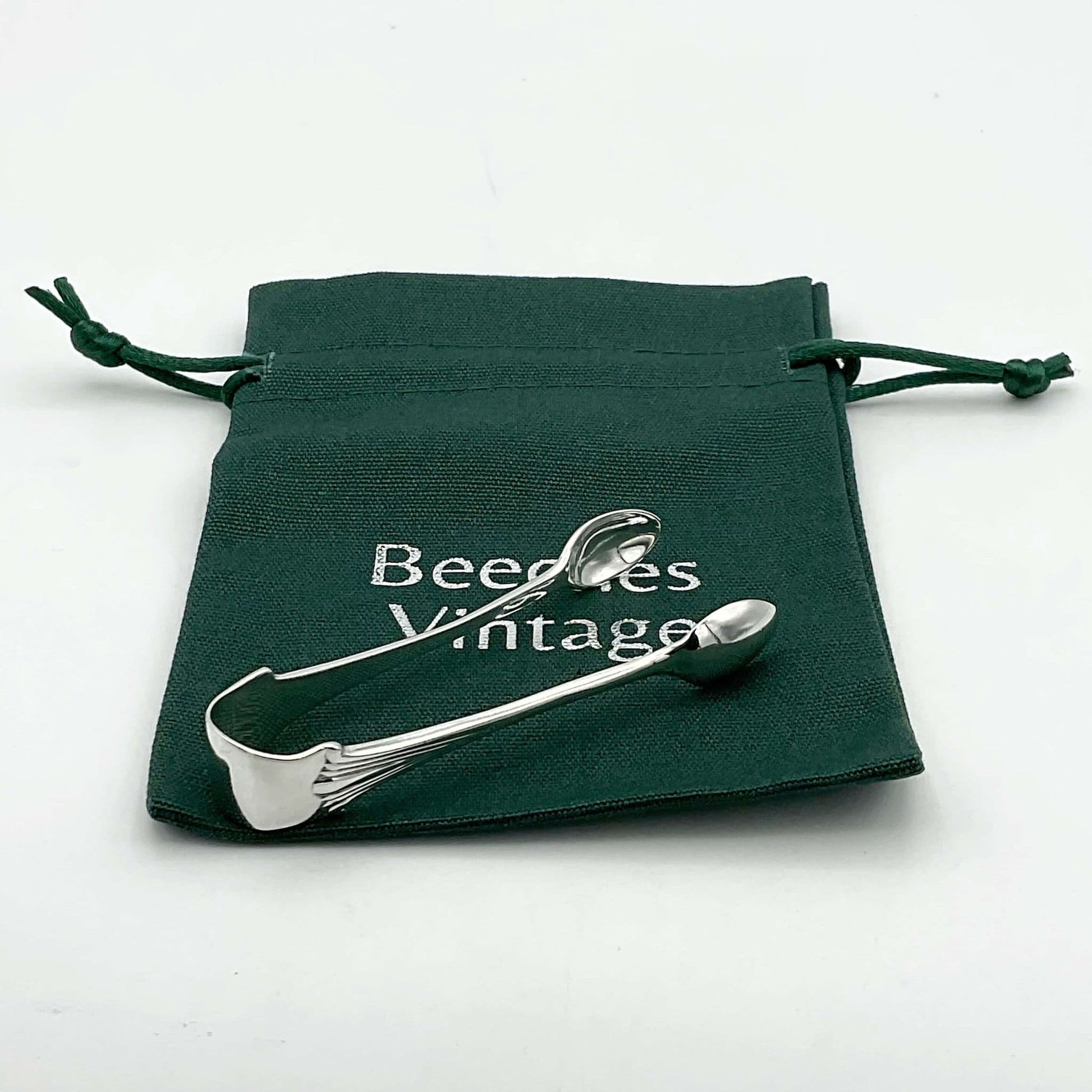 Silver sugar tongs showing an art deco design to the art on a white background sitting on a green Beeches Vintage bag