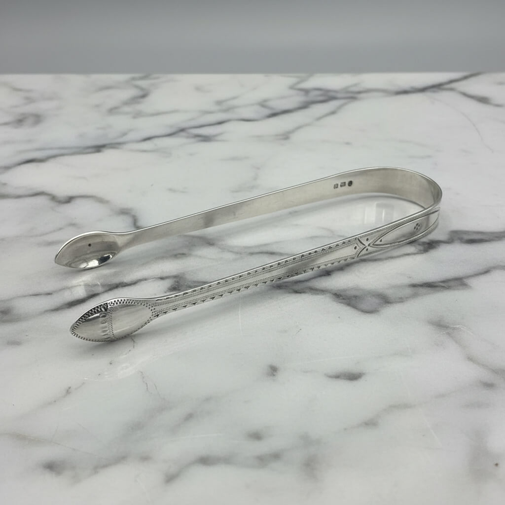 side view of antique silver sugar tongs with engraving on a marble surface