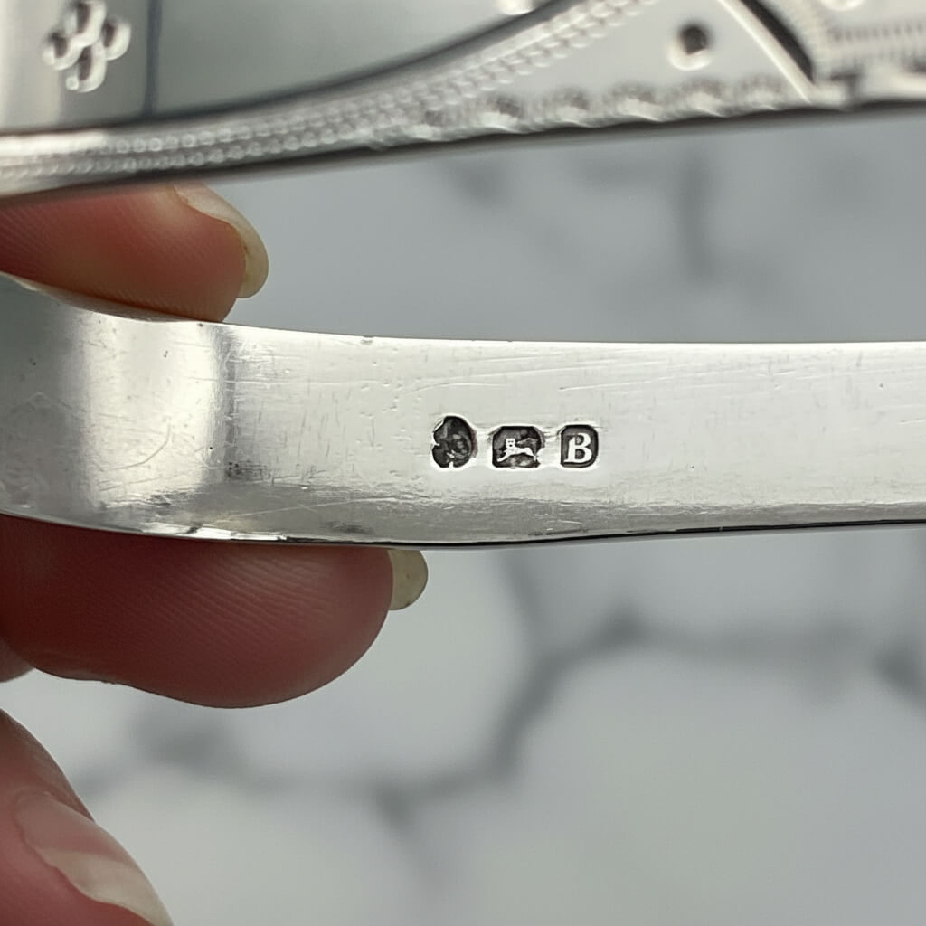 hallmarks on the inside of antique silver sugar tongs held in fingers with a marble background